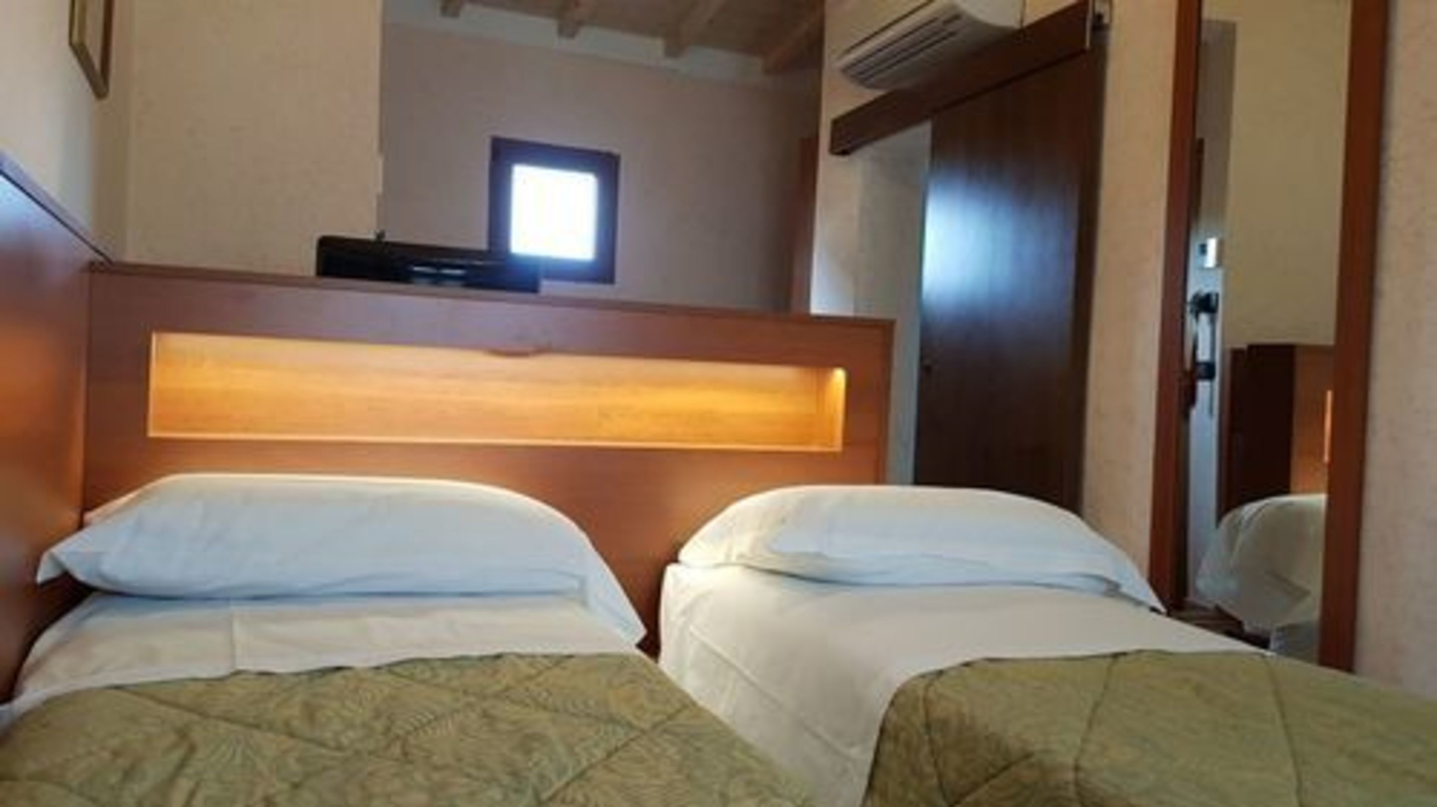 Camera d'albergo con due letti, una testiera in legno, un piccolo finestrino e un condizionatore d'aria.