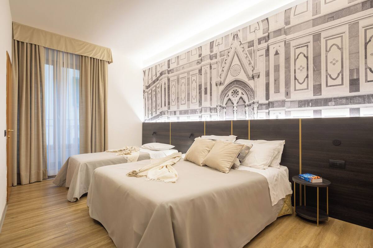 A hotel room with two beds, a large tapestry on the wall, a small side table, and a window with curtains.