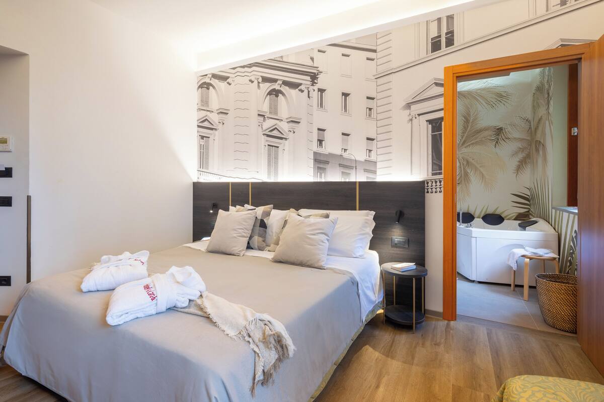 A modern hotel room with a large bed, a small bedside table, a bathroom with a bathtub, and a view of a cityscape through a glass door.