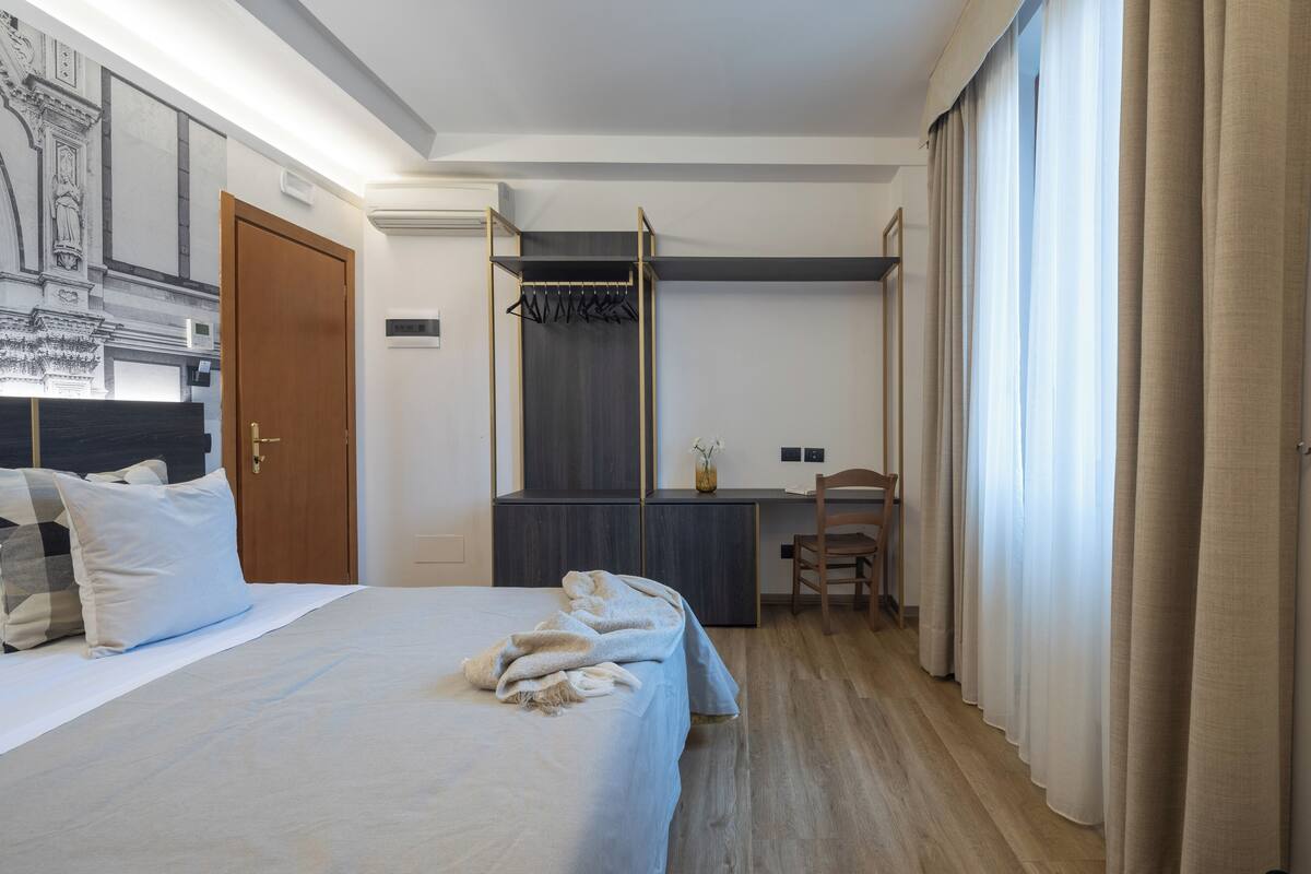 A hotel room with a bed, a desk, and a chair. There is a wardrobe and a window with curtains.