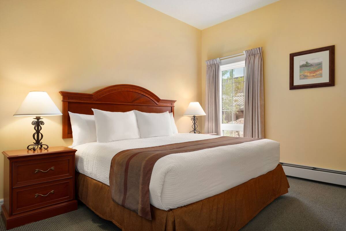 A hotel room with a large bed, two bedside tables with lamps, a window with curtains, and a framed picture on the wall.