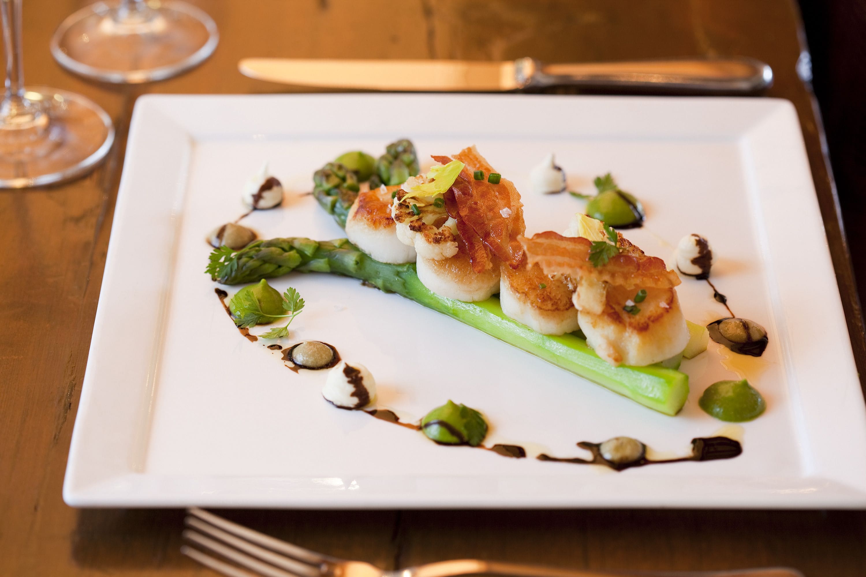 Une assiette carrée blanche sur laquelle sont disposés desSaint-Jacques poêlés, des asperges et diverses garnitures, le tout présenté sur une table en bois.