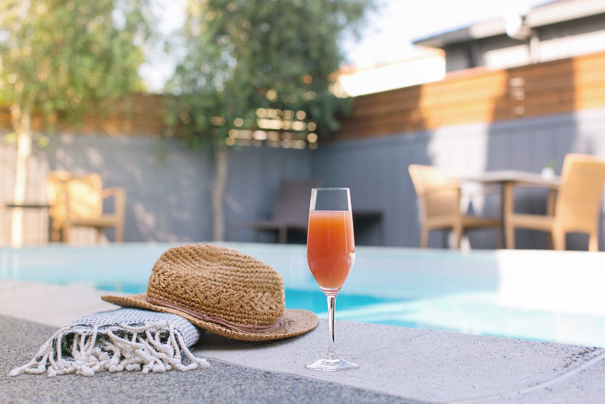 A poolside setting with a clear glass of a pinkish drink, a straw hat, and a woven bag on the edge of the pool.