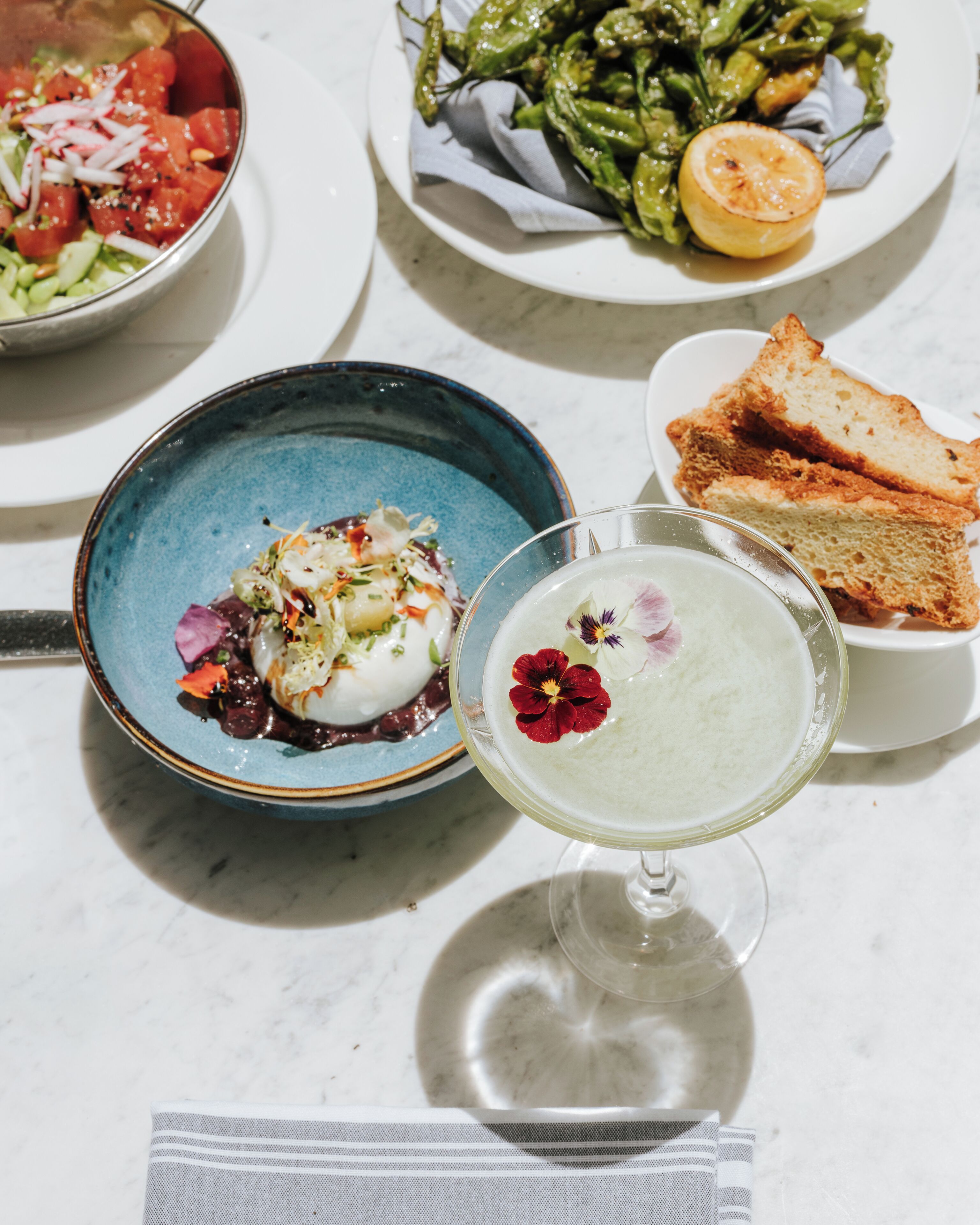 Une table où l’on trouve une variété de plats, notamment une salade, une assiette de légumes grillés, un dessert et un cocktail.