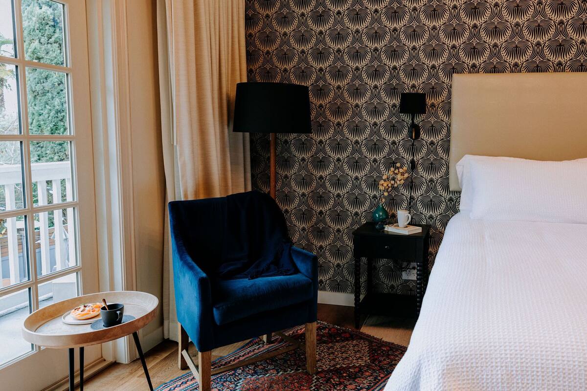 A hotel room with a bed, a blue armchair, a small table with a plate of food, a lamp, and a patterned wallpaper.