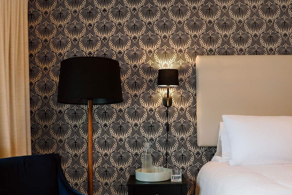 A hotel room with a patterned wallpaper, a blue armchair, a bedside table with a lamp, and a bed with white linens.