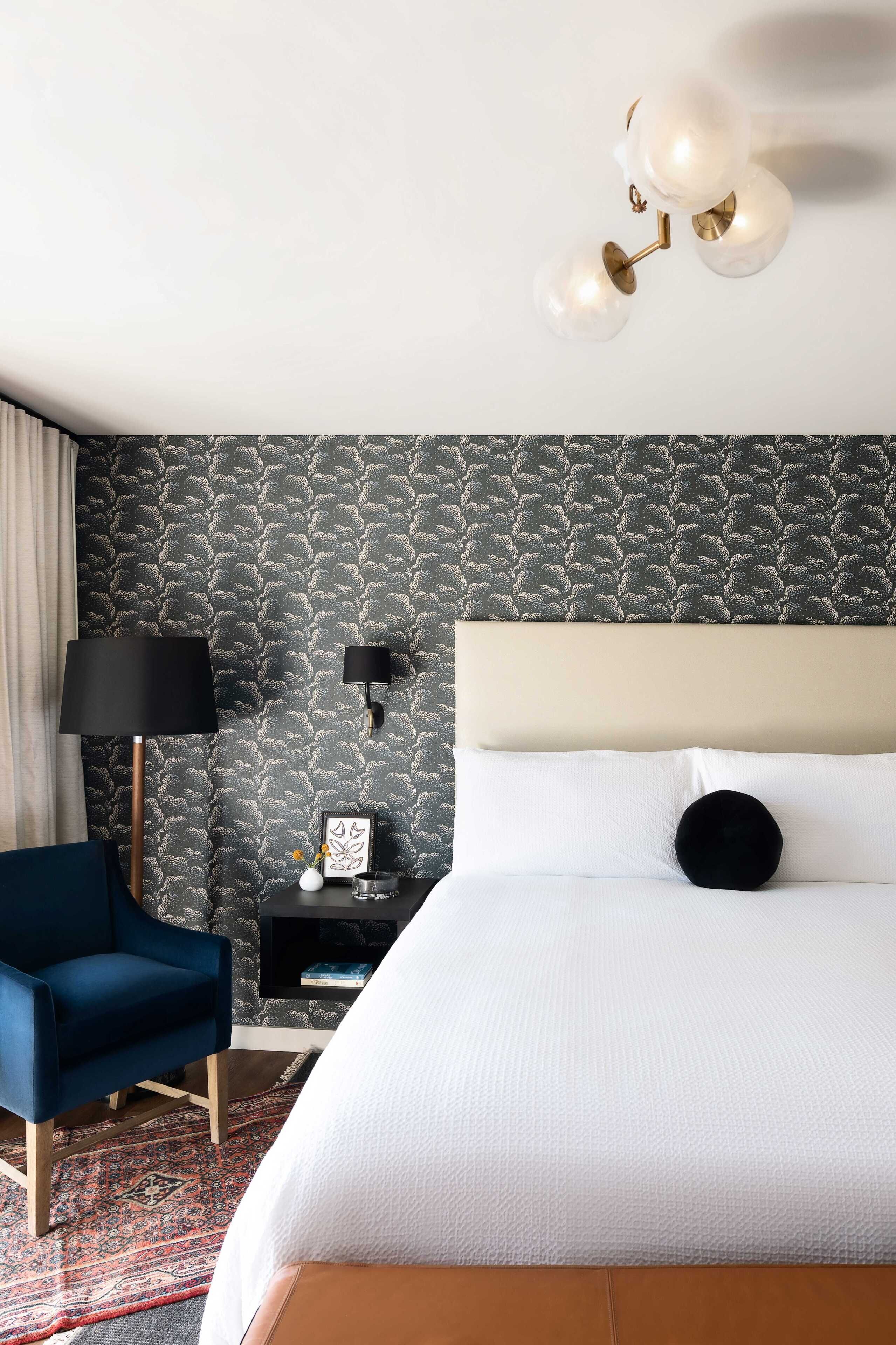 Una camera d'albergo con un letto, una poltrona blu, un comodino, una lampada e una parete decorata.