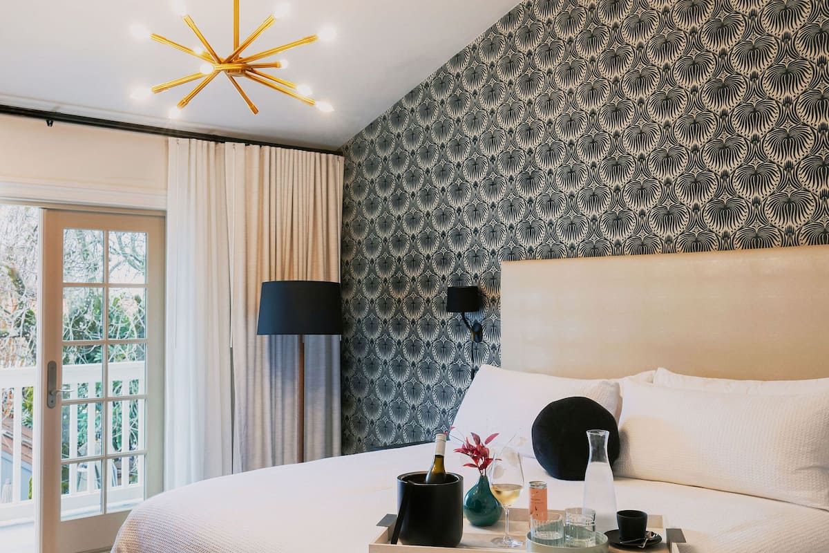 A modern bedroom with a large bed, a ceiling light fixture, a patterned wall, and a view through a glass door.