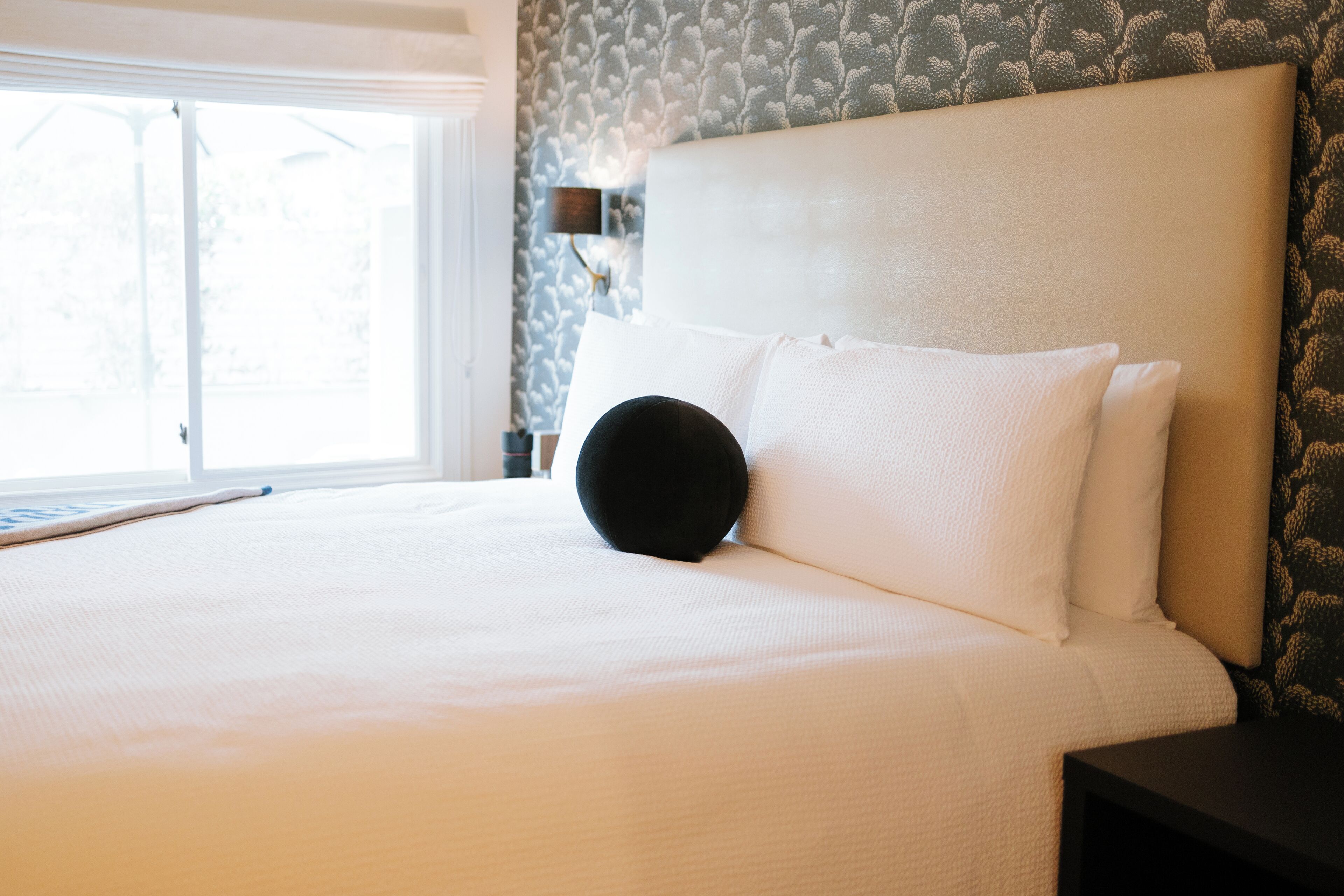 A neatly made bed with white linens and a black pillow, a bedside table, and a wall with a patterned wallpaper.
