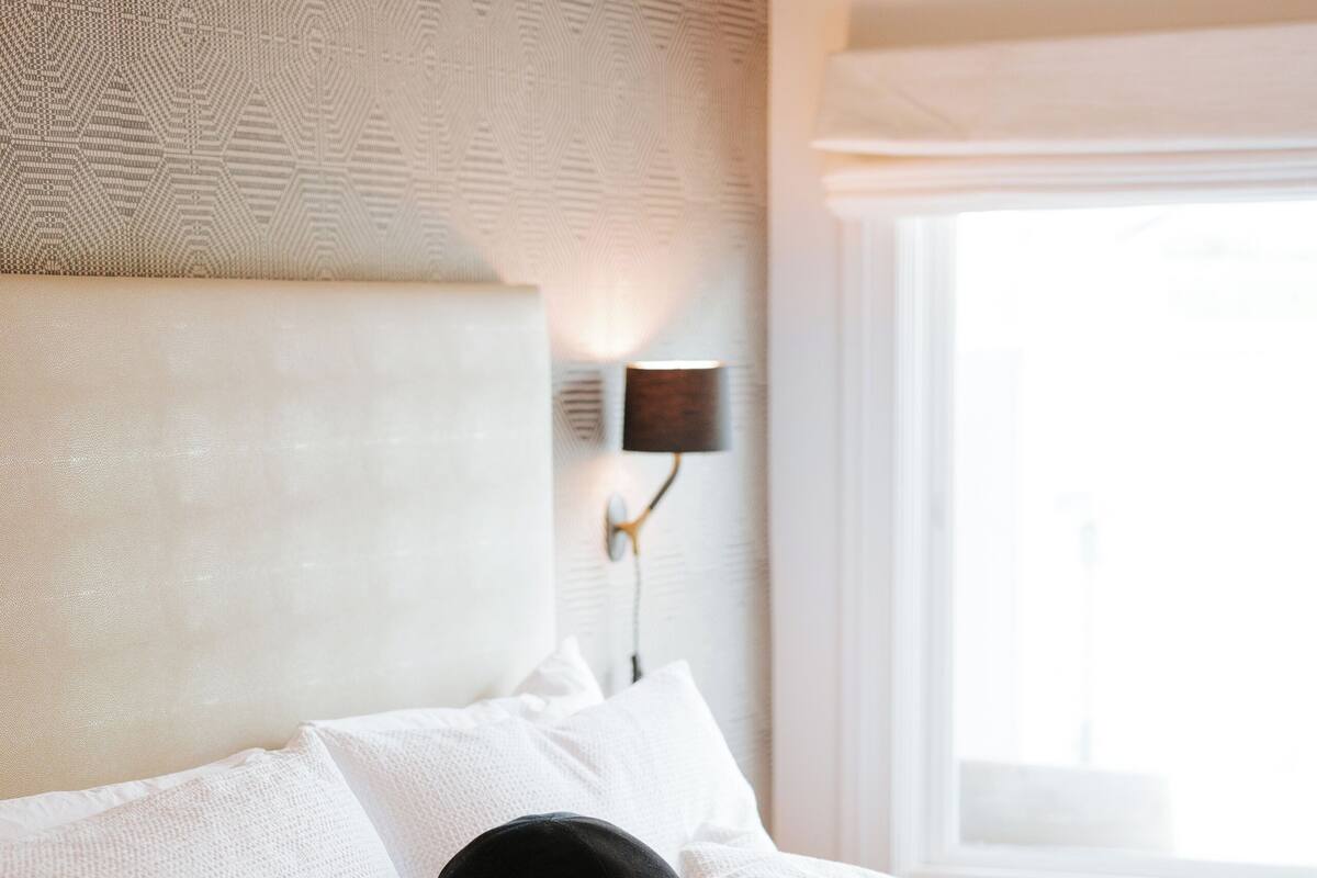 A neatly made bed with white bedding and a black pillow, a wall-mounted light fixture, and a window with daylight.