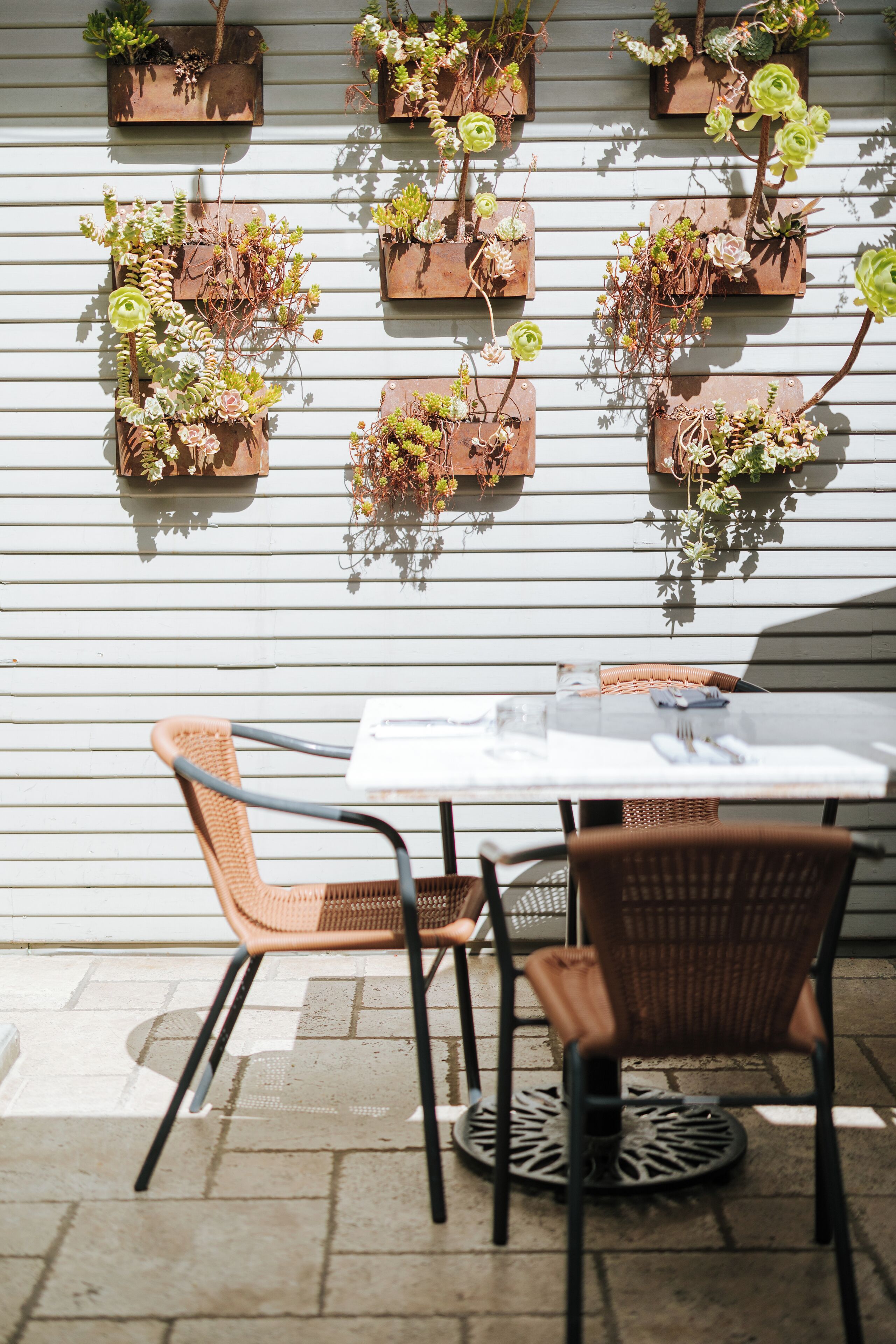 A patio area with a table and chairs, a white wall with wooden planters, and a brick floor.
