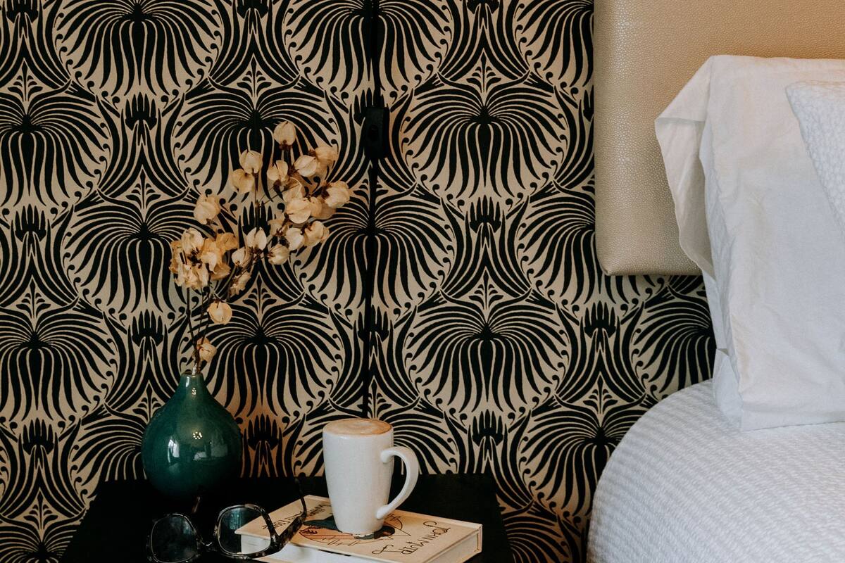 A neatly made bed with a white comforter, a black nightstand with a green vase and a white mug, and a wall with a black and beige patterned wallpaper.