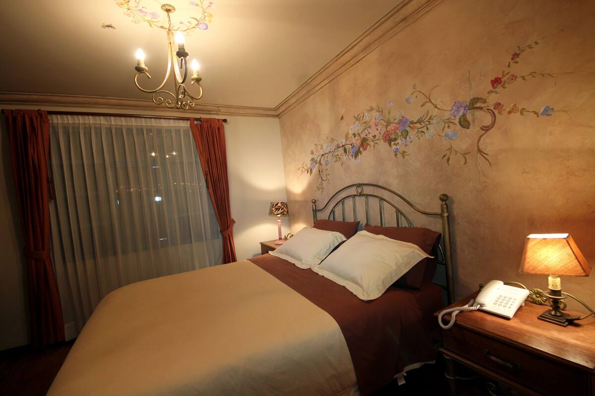 A hotel room with a large bed, a bedside telephone, a lamp, and a floral mural on the wall.