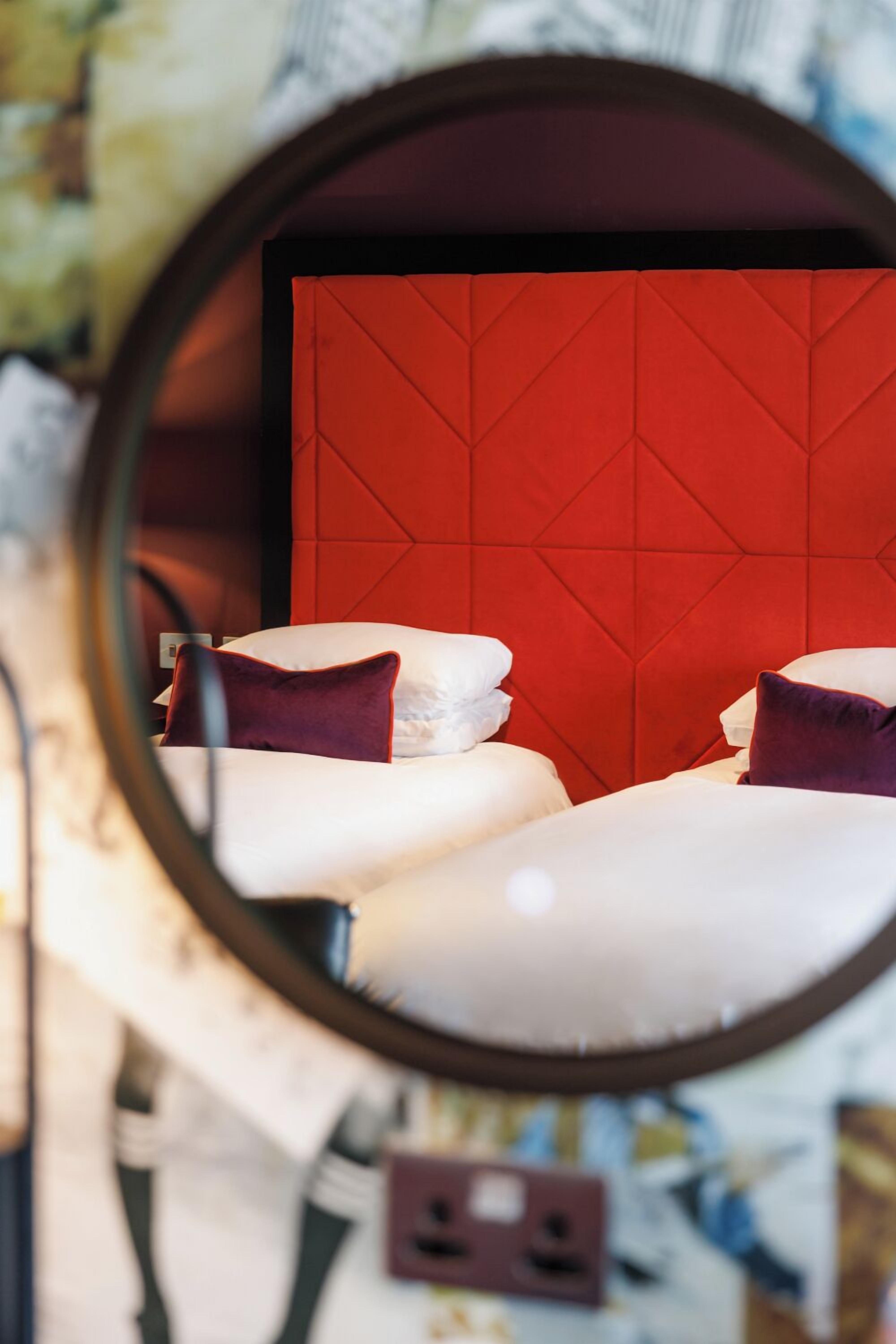 Une chambre d’hôtel avec une tête de lit rouge, du linge de lit blanc et un miroir rond qui reflète la pièce.