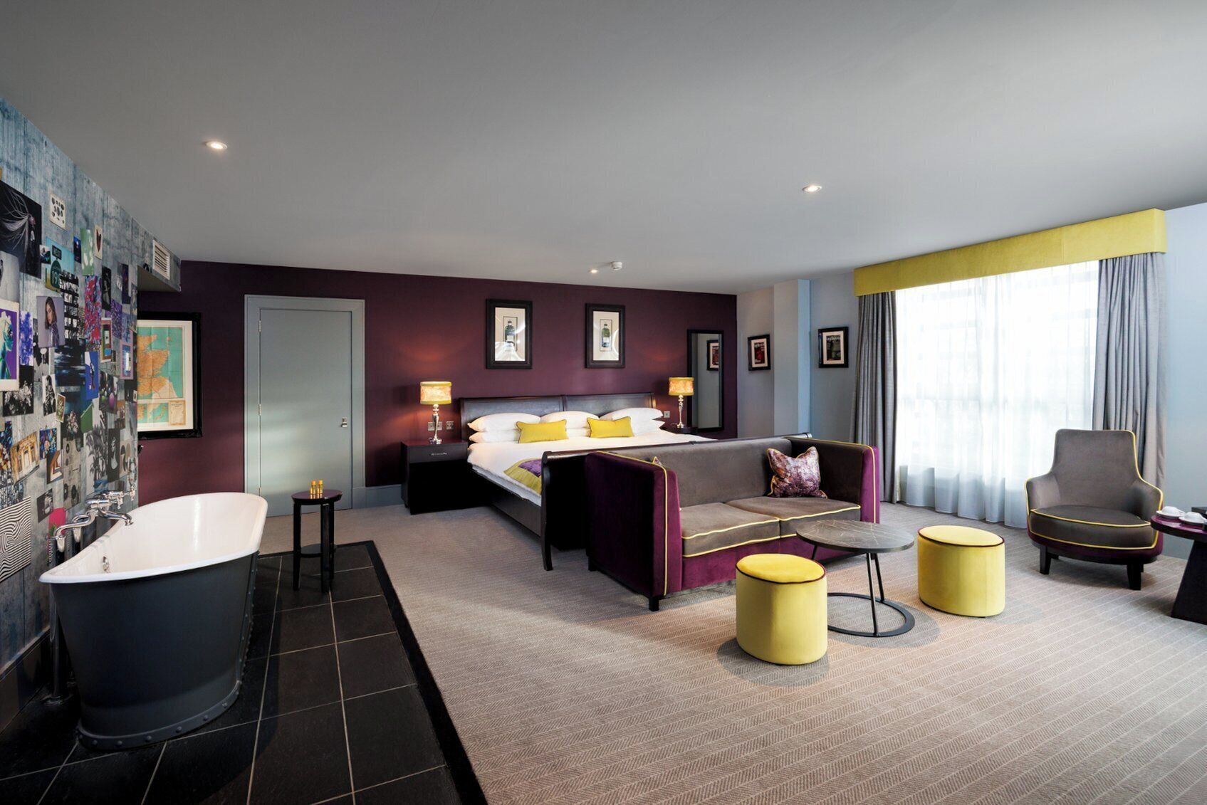 A modern hotel room with a large bed, a sitting area with a sofa and armchairs, a bathtub, and a large window with curtains.