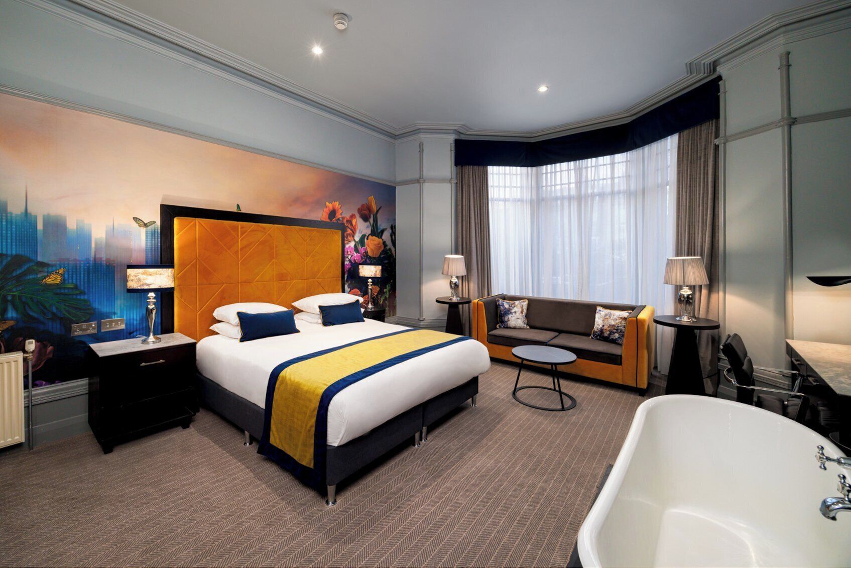 A hotel room with a large bed, a sofa, a small round table, a bathtub, and a cityscape mural on the wall.