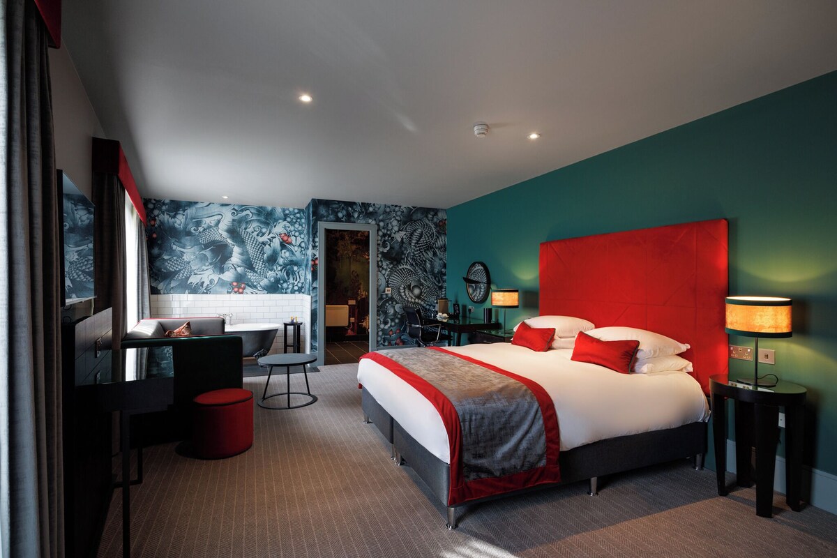 A modern hotel room with a large bed, a desk, a chair, a red accent wall, and a bathroom visible through an open door.