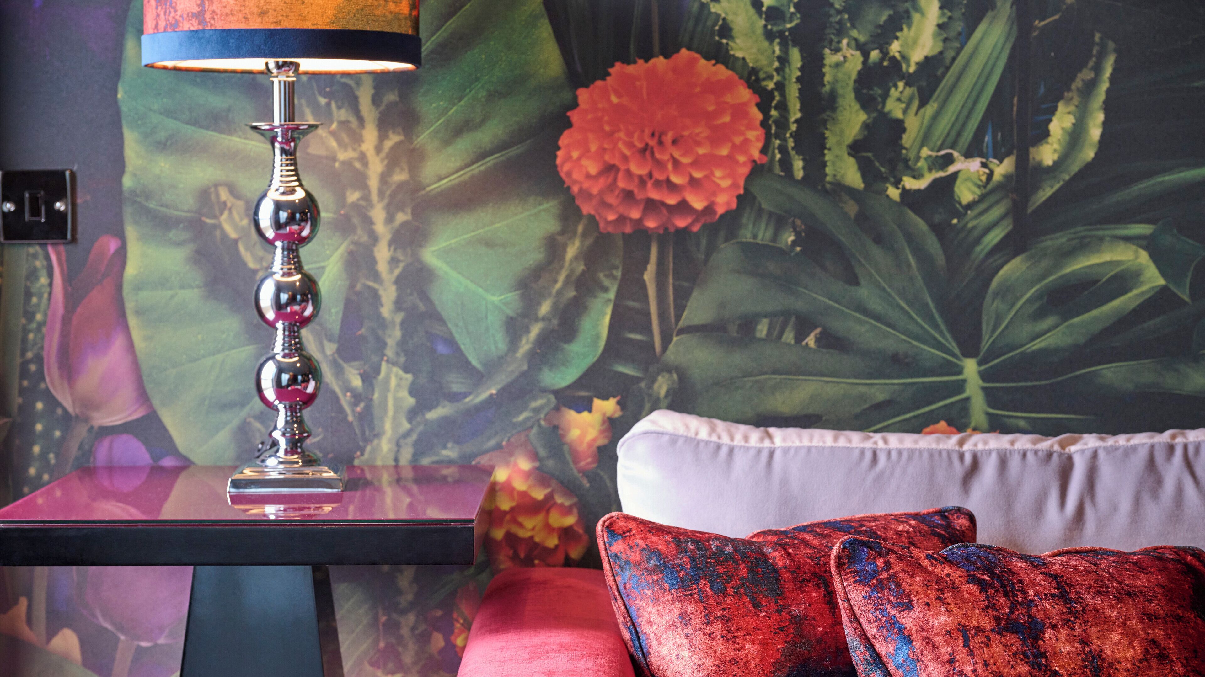 A room with a modern lamp on a side table, a sofa with a patterned throw pillow, and a vibrant floral mural on the wall.