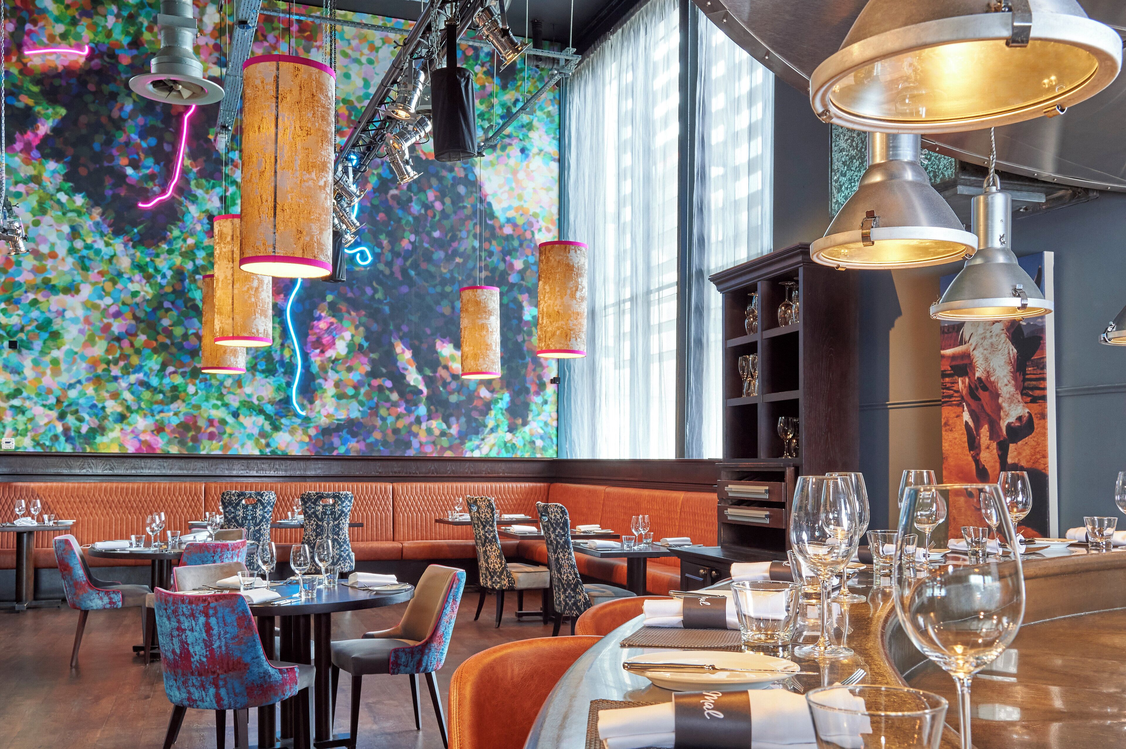 Een restaurant met een kleurrijke abstracte muurschildering, moderne hanglampen en diverse zitplaatsen, waaronder hoekbankjes en tafels.