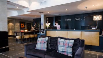 A modern hotel lobby with a reception desk, a bar area, and a seating area with a dark-colored sofa and plaid cushions.