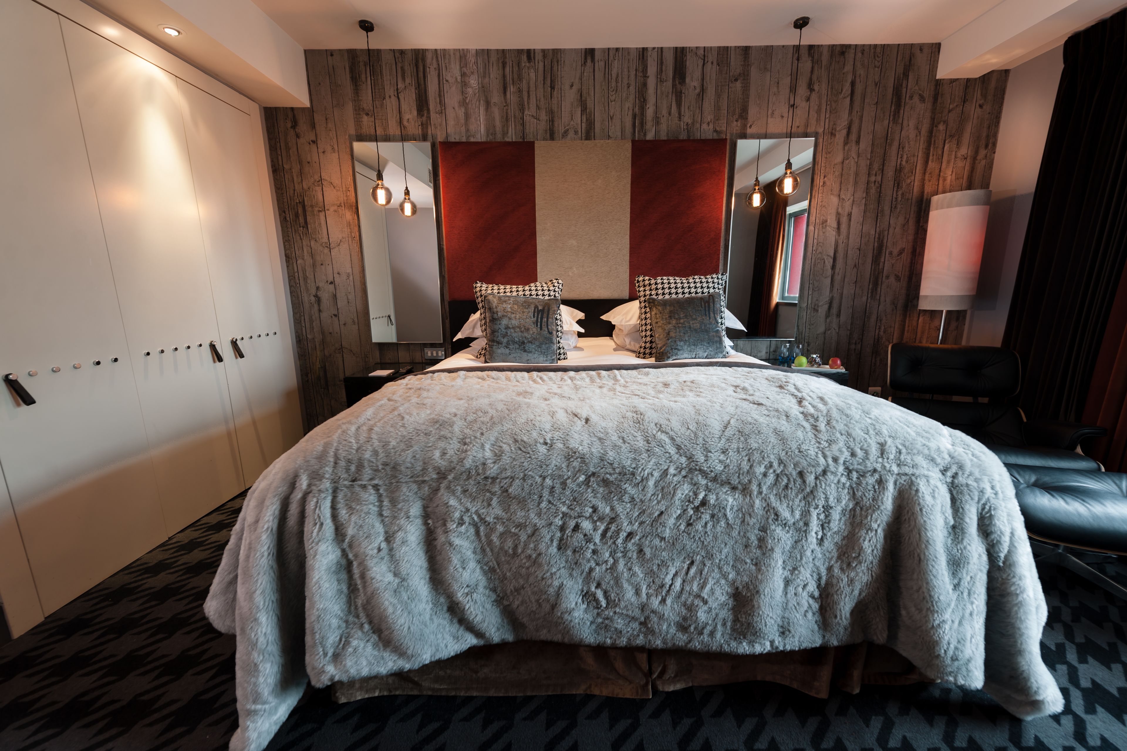 A modern hotel room with a large bed, a wardrobe, a mirror, and a desk with a lamp.