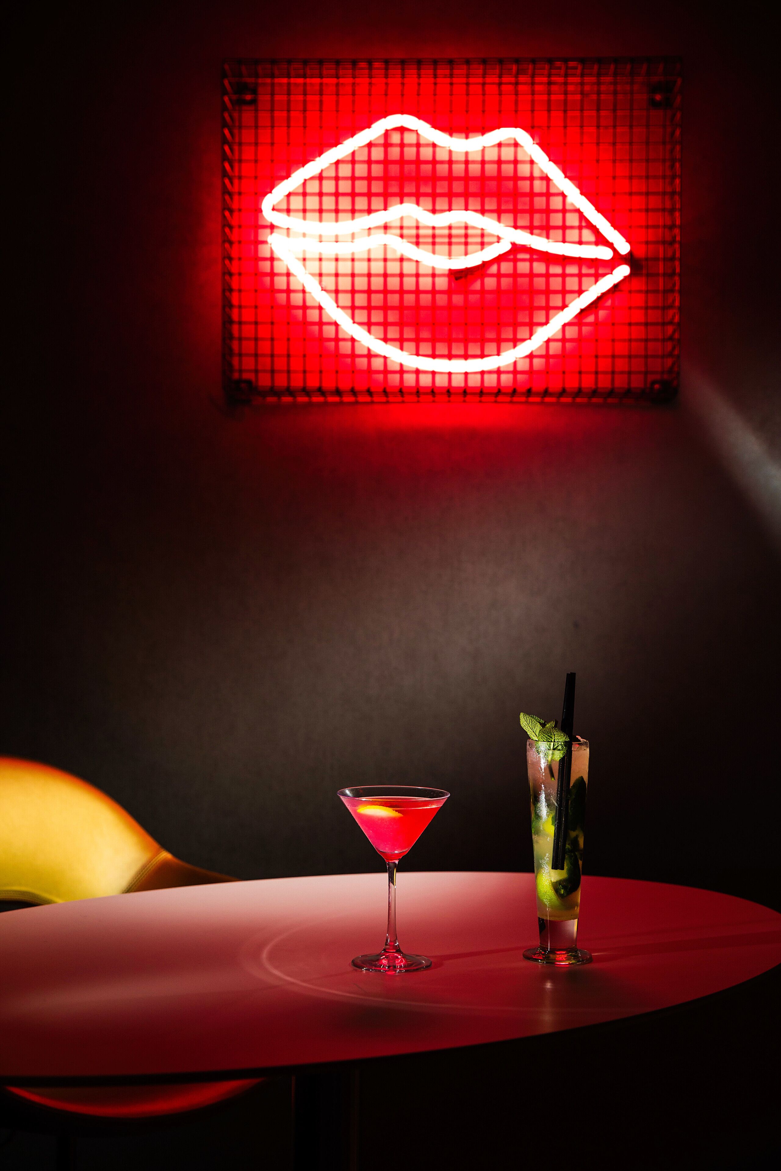 Eine Barbereich mit einem Neon-Lippenschild, einem Martini-Glas und einem hohen Glas mit Trinkhalm und Minze auf einem roten Tisch.