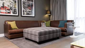 A modern living room with a brown leather sofa, a grey ottoman, and a small side table with a lamp. There are two framed pictures on the wall and a window with curtains.