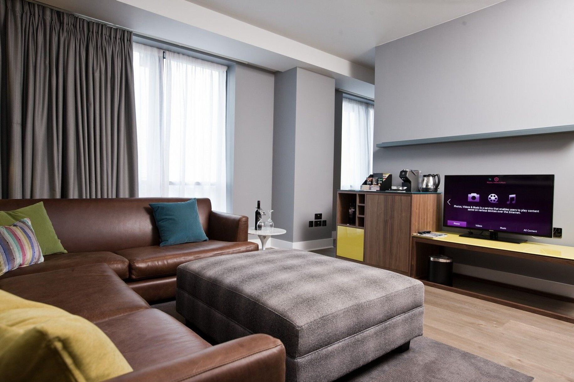 Moderni hotellihuone, jossa on nahkasohva, harmaa rahi, pieni pyöreä pöytä ja televisio puisen jalustan päällä.