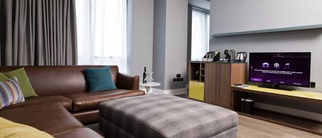 A modern hotel room with a leather sofa, a grey ottoman, a small round table, and a TV on a wooden stand.