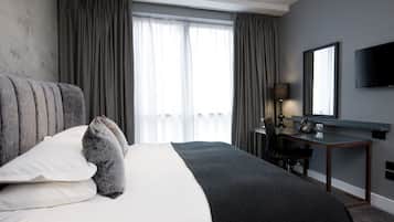 A hotel room with a large bed, a desk with a lamp, a telephone, and a flat-screen TV on the wall.