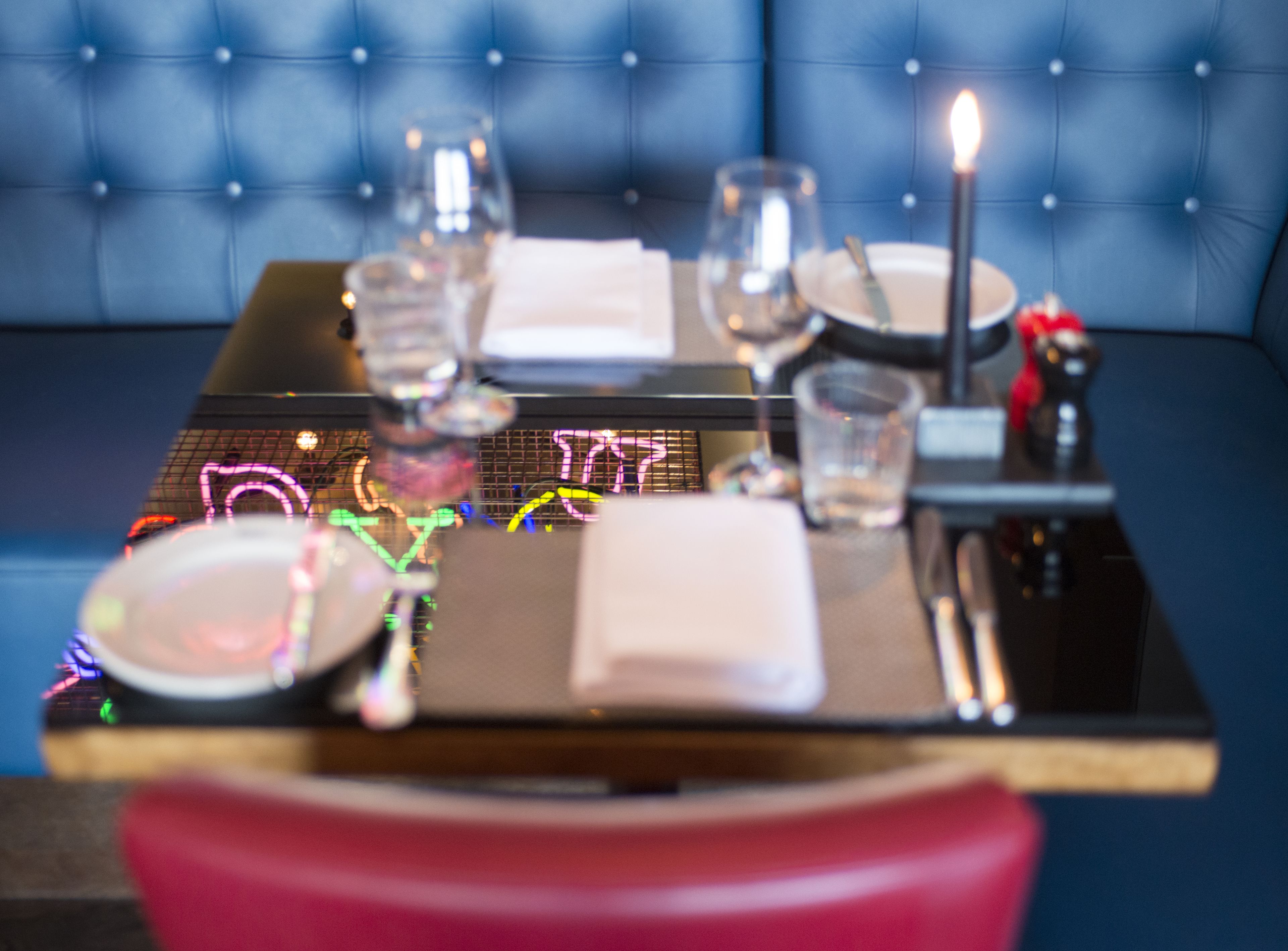A dining table set with plates, glasses, and a candle, with a neon sign in the background.