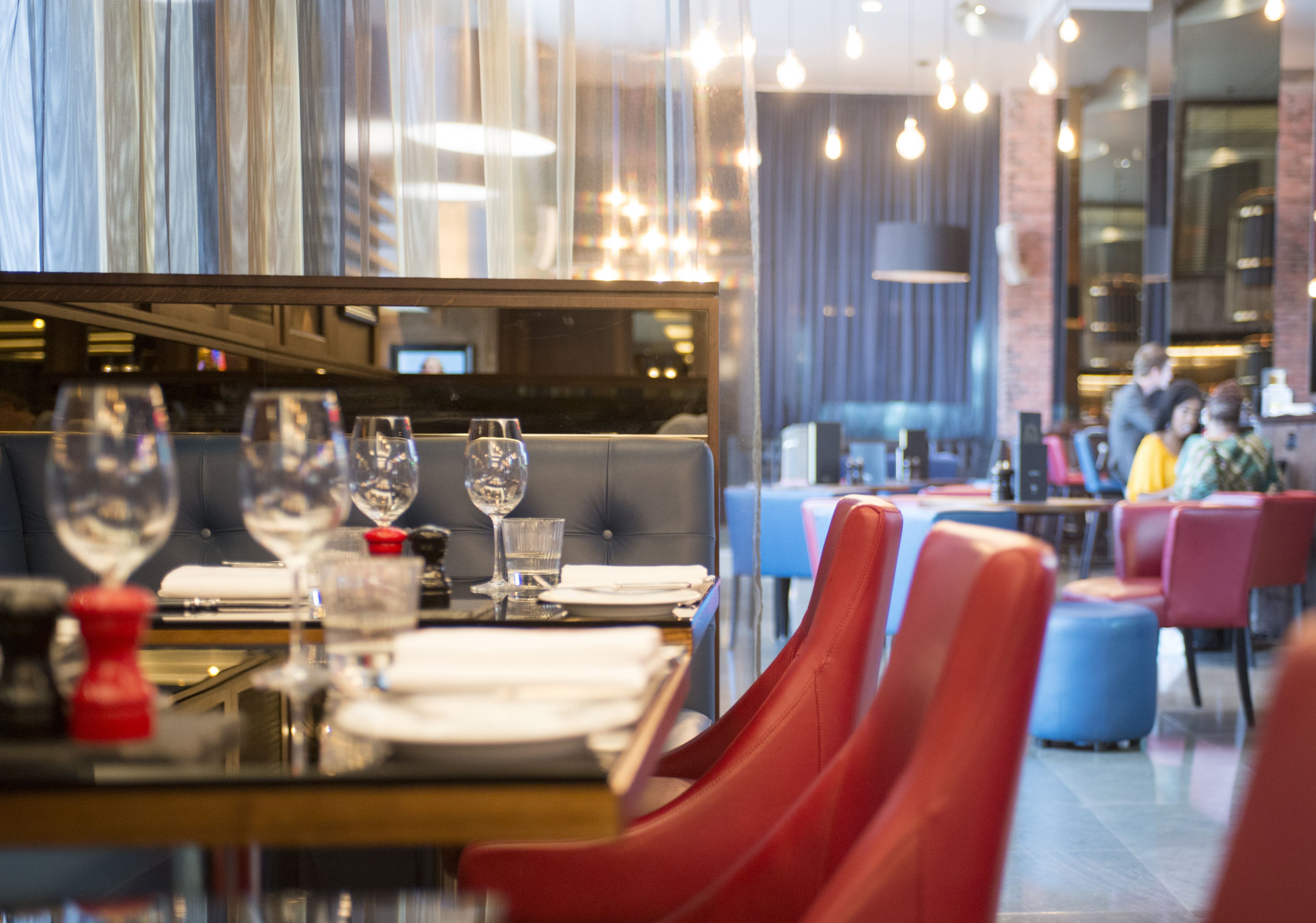 A restaurant with red upholstered chairs, a glass table with wine glasses and water glasses, and a modern interior with hanging lights and a brick wall.