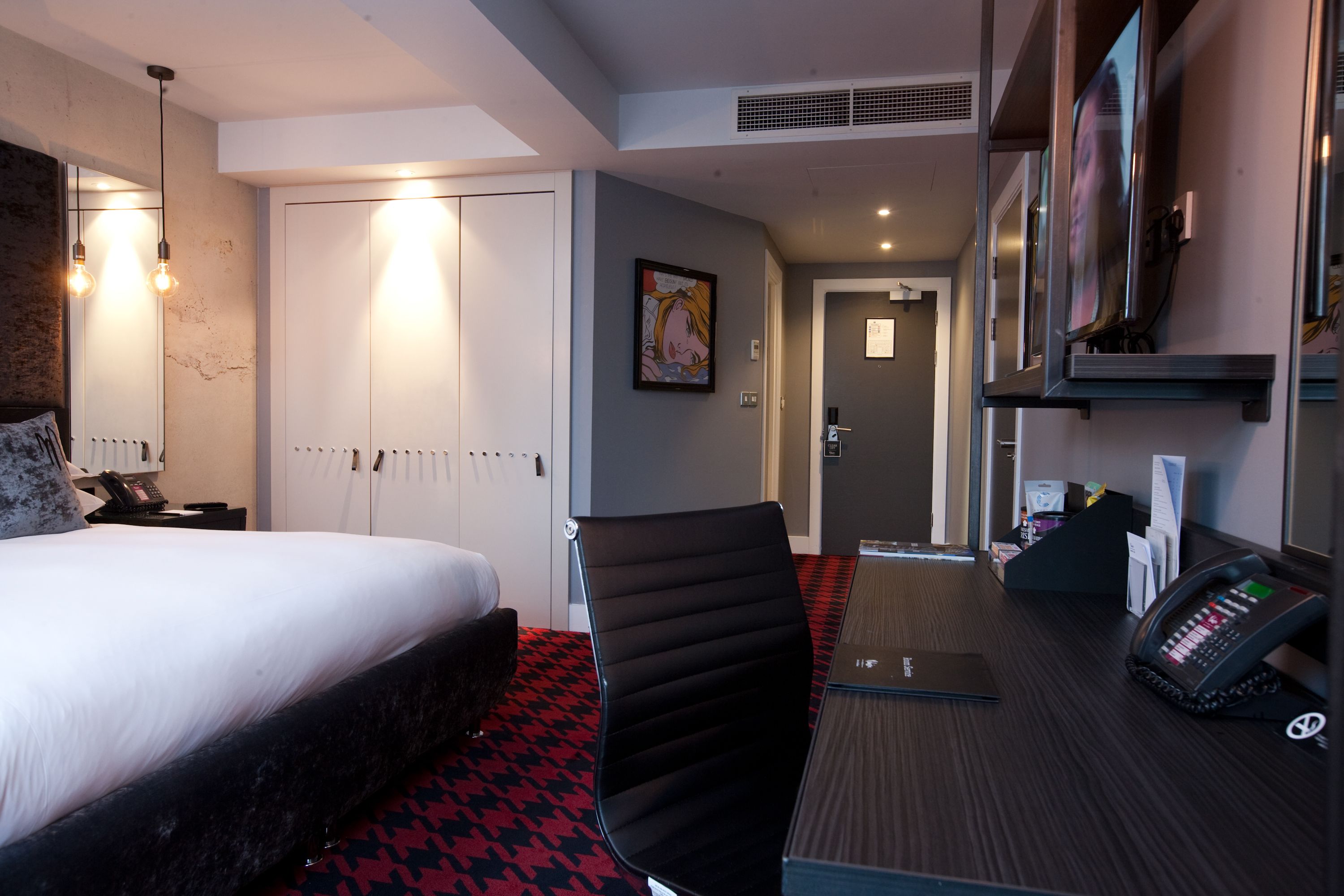 Quarto de hotel com cama, secretária com telefone, uma cadeira e porta para o banheiro.