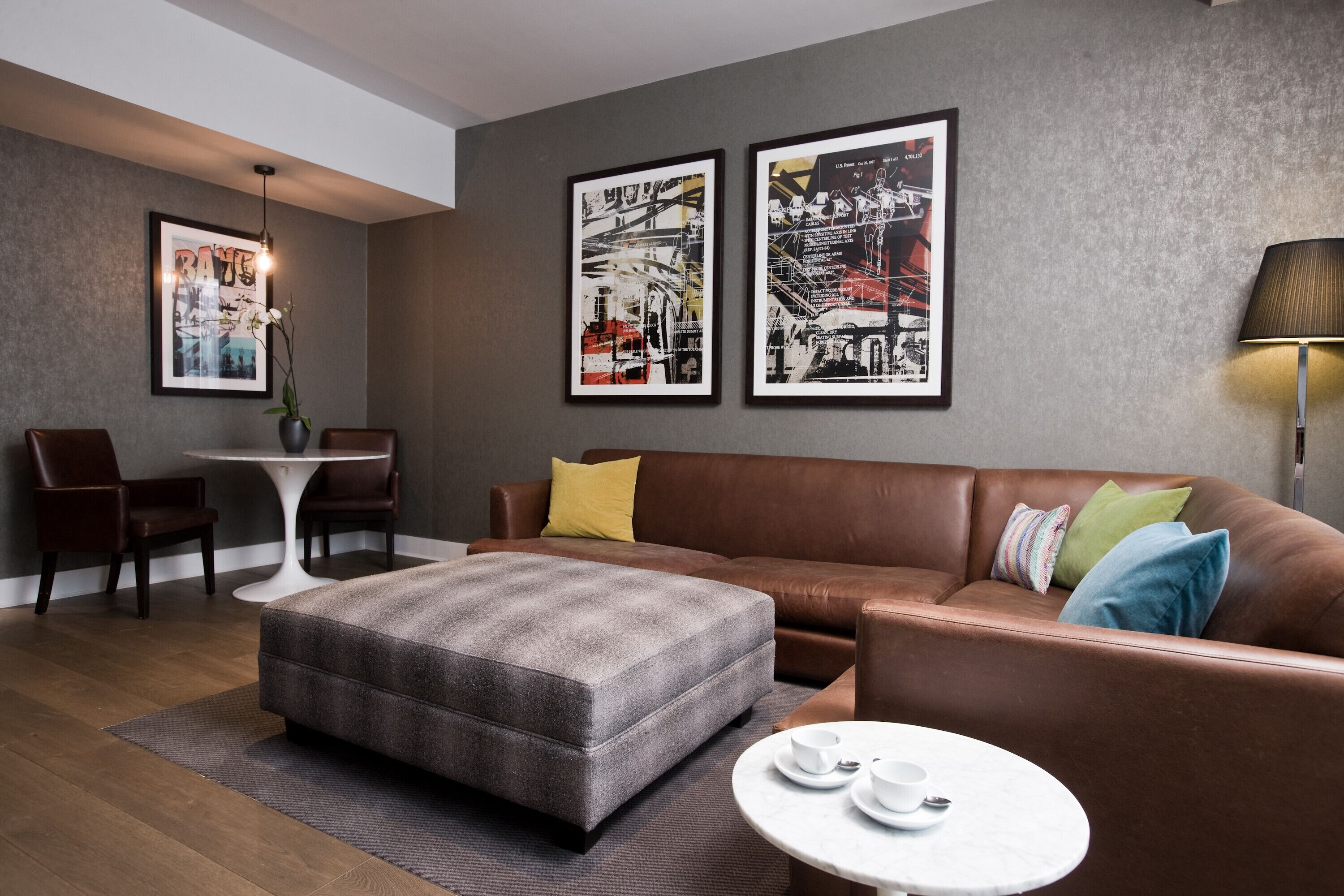 A modern living room with a brown leather sofa, a grey ottoman, a white coffee table, and two framed abstract artworks on the wall.