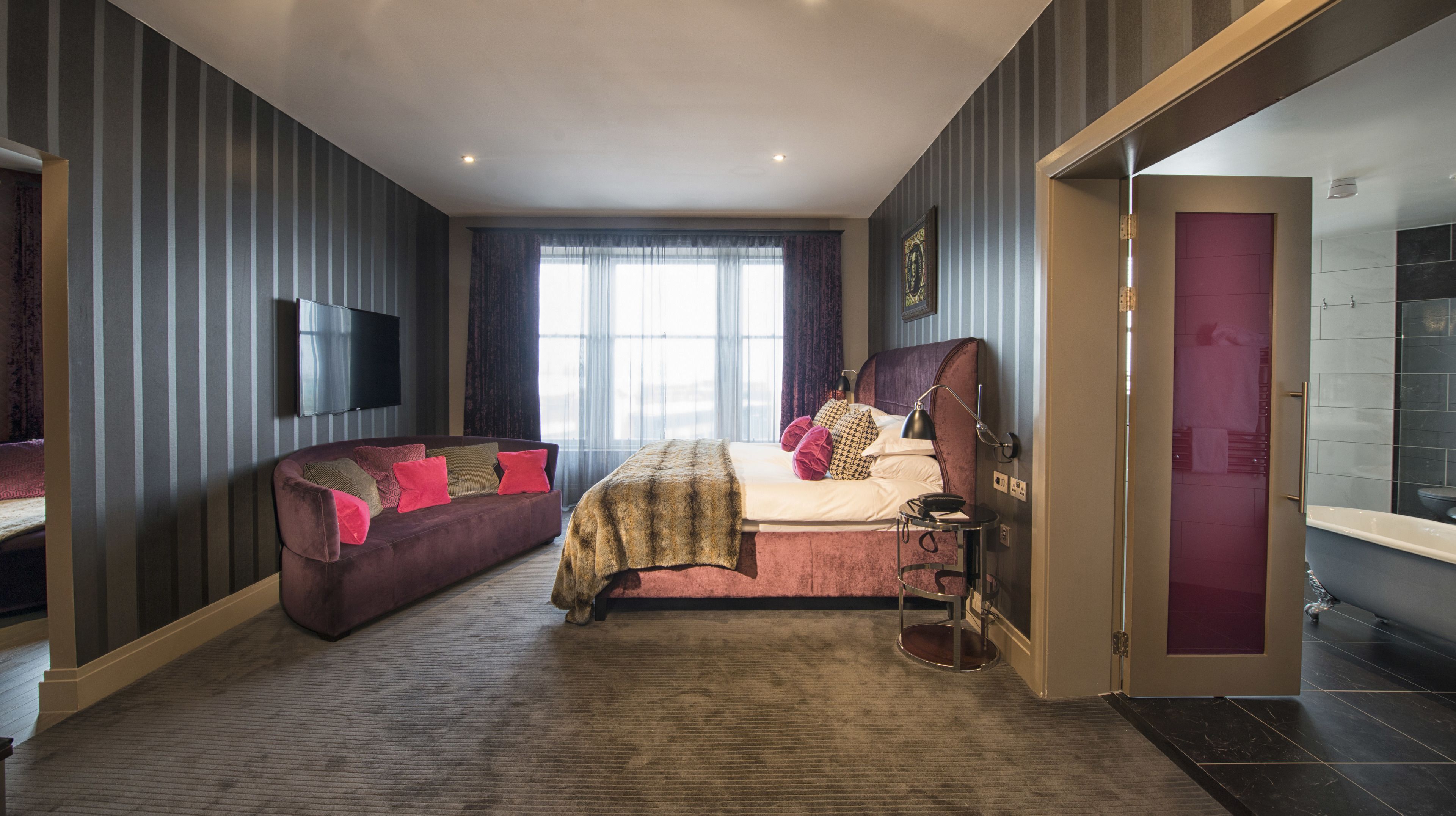 A modern bedroom with a large bed, a sofa, a TV, and a bathroom visible through an open door.