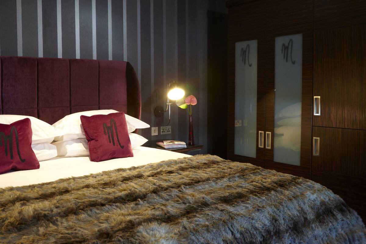 Ein ordentlich bezogenes Bett mit einem weichen, roten Kopfteil, einer flauschigen, grauen Decke und zwei roten Kissen mit einem Monogramm.