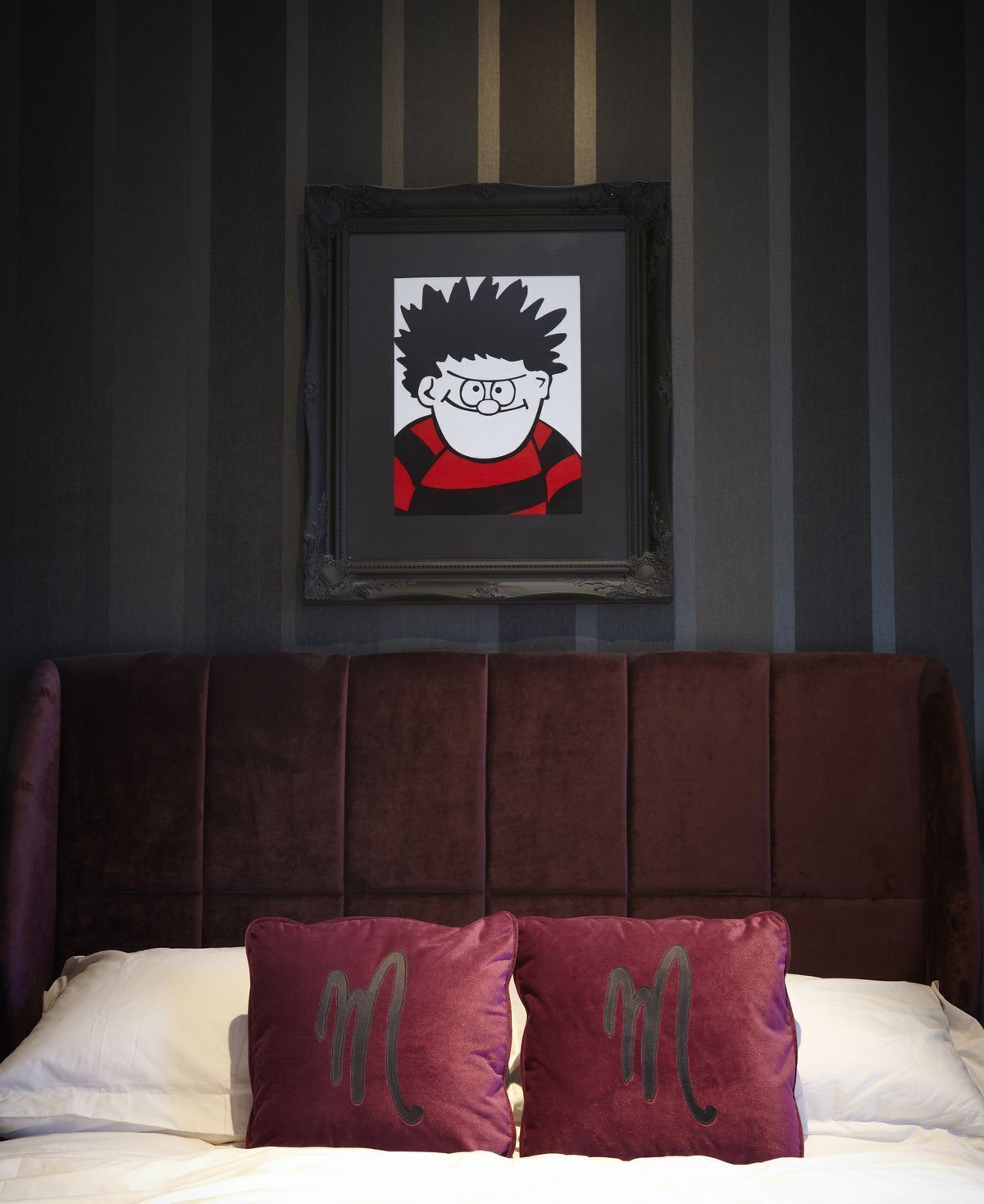 A bedroom with a dark brown headboard, a framed cartoon character on the wall, and two pillows with a 'm' design.