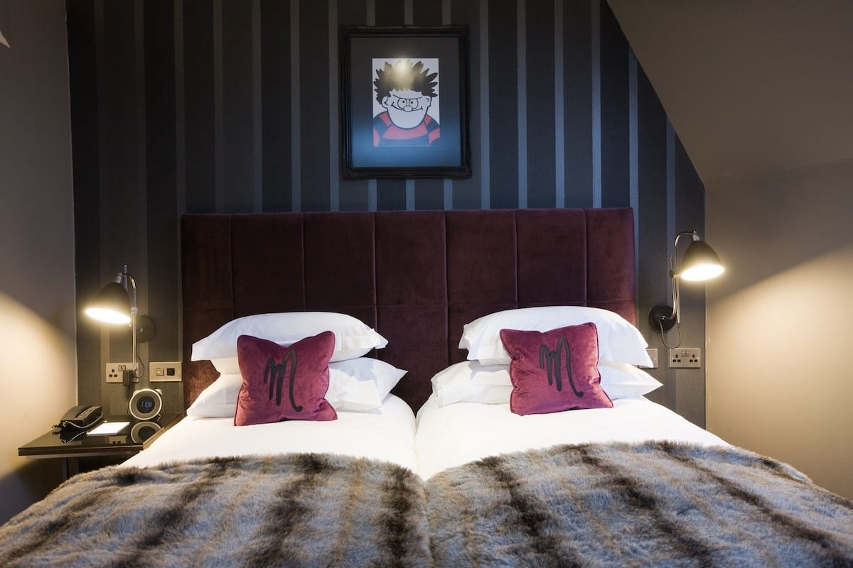 Ein Hotelzimmer mit einem großen Bett, zwei mit Monogramm versehenen Kissen, einem Kopfteil, Nachttischlampen, einer Uhr und einem gerahmten Bild an der Wand.