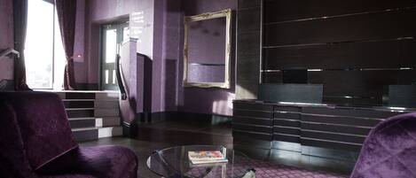 A lobby with purple velvet chairs, a glass coffee table, a framed mirror, and a staircase.