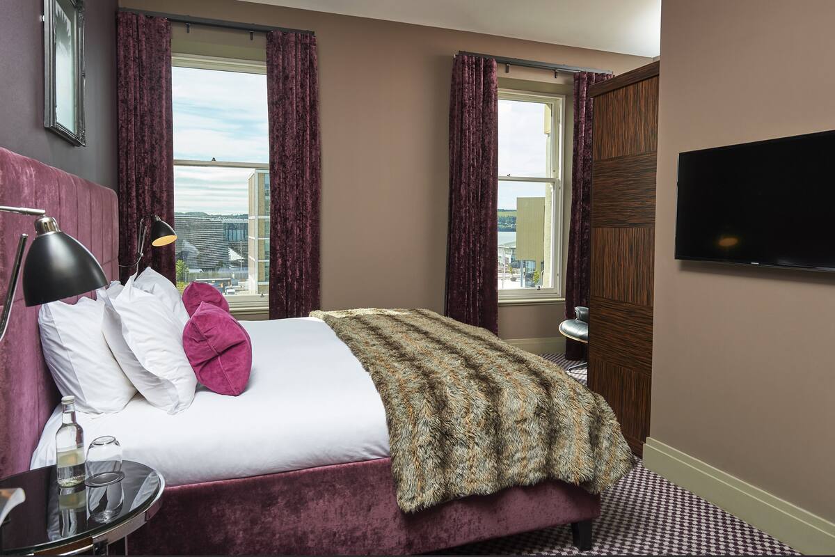 Ein Hotelzimmer mit einem großen Bett, einem Fernseher, einem Nachttisch mit Lampe und einem Fenster mit Vorhängen.