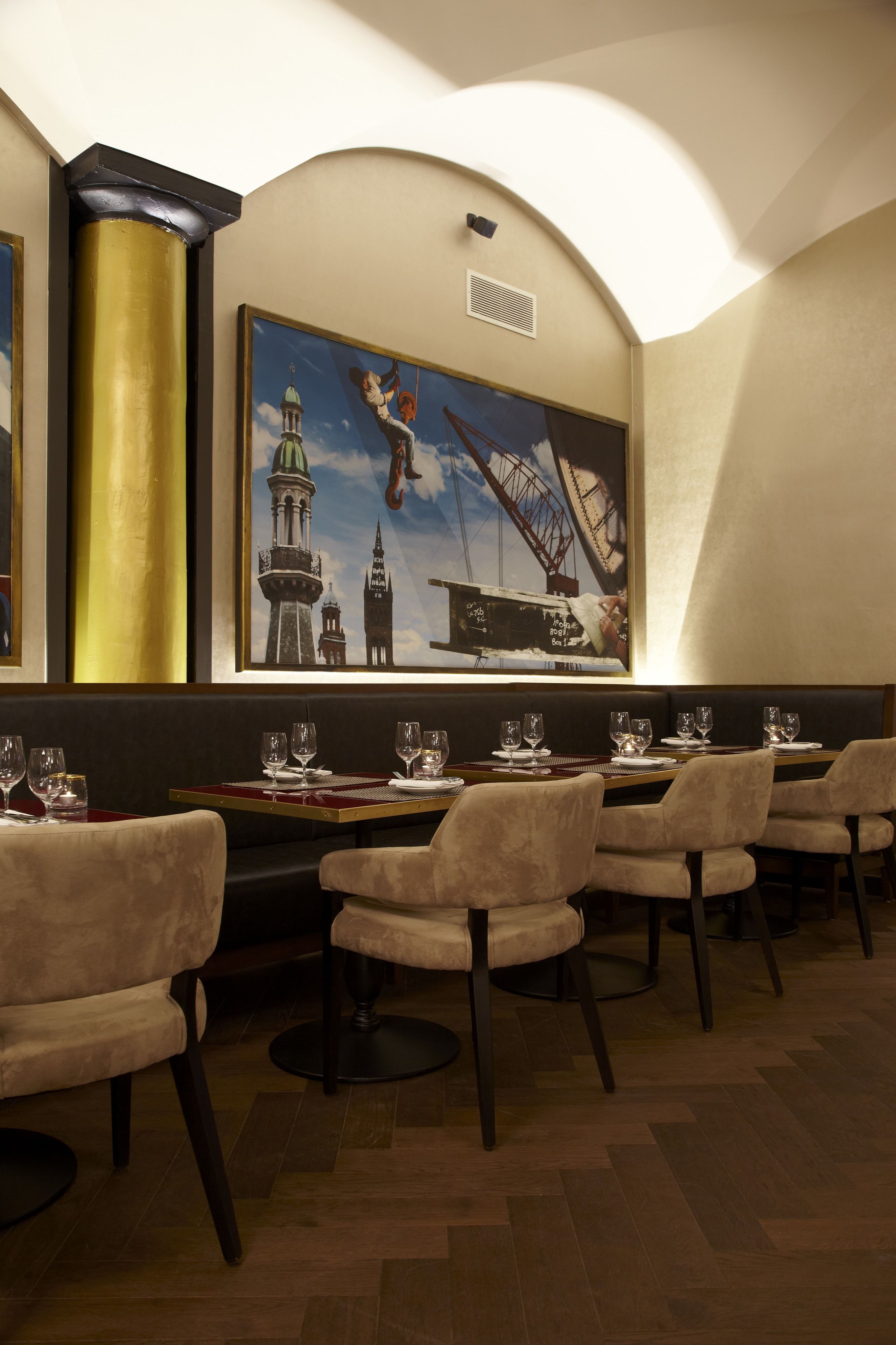 A restaurant with a high ceiling, a large painting of a bridge and tower, and a row of tables with chairs.