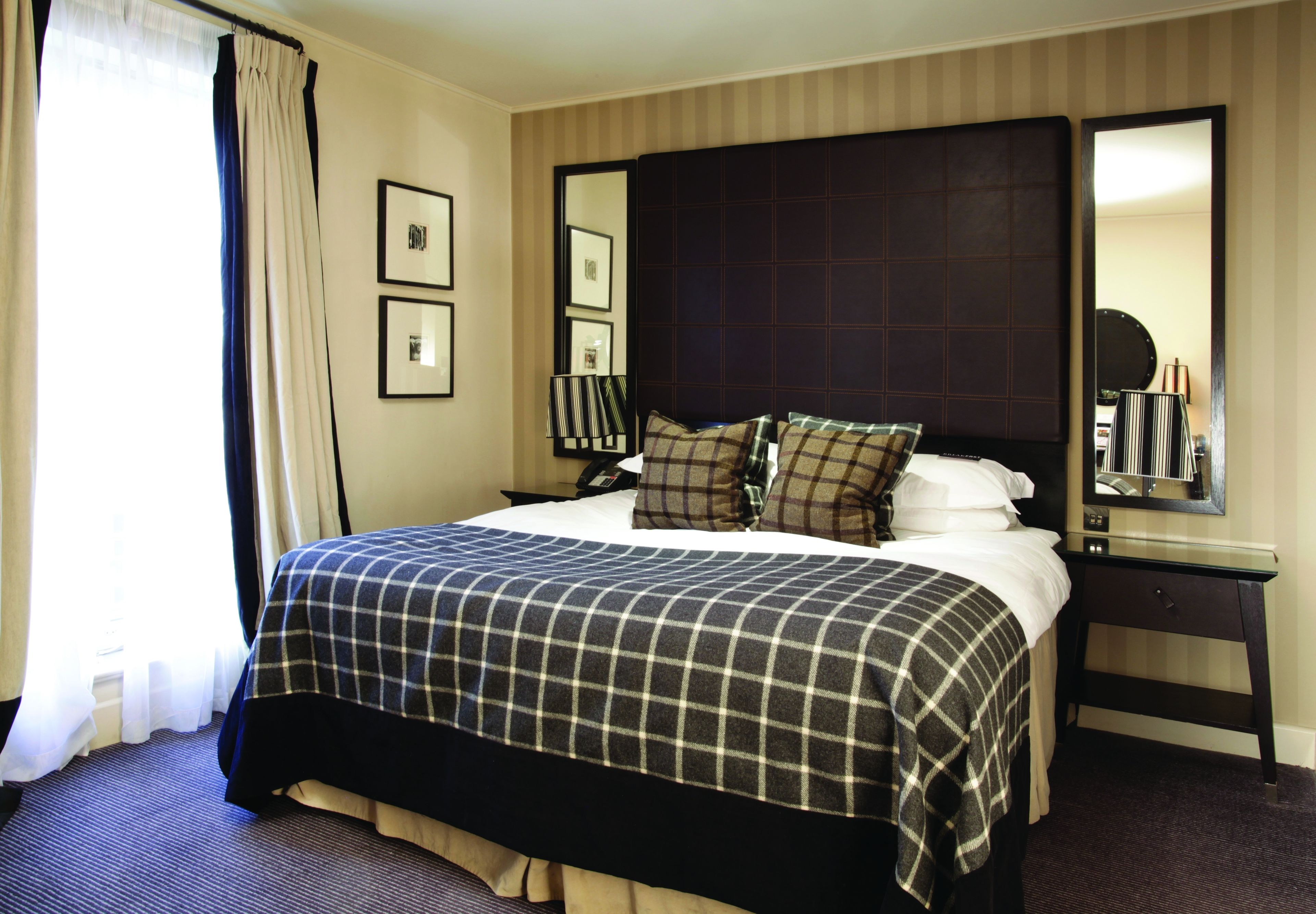 Een hotelkamer met een groot bed, een donker hoofdeinde, een nachtkastje met een spiegel en ingelijste foto's aan de muur.