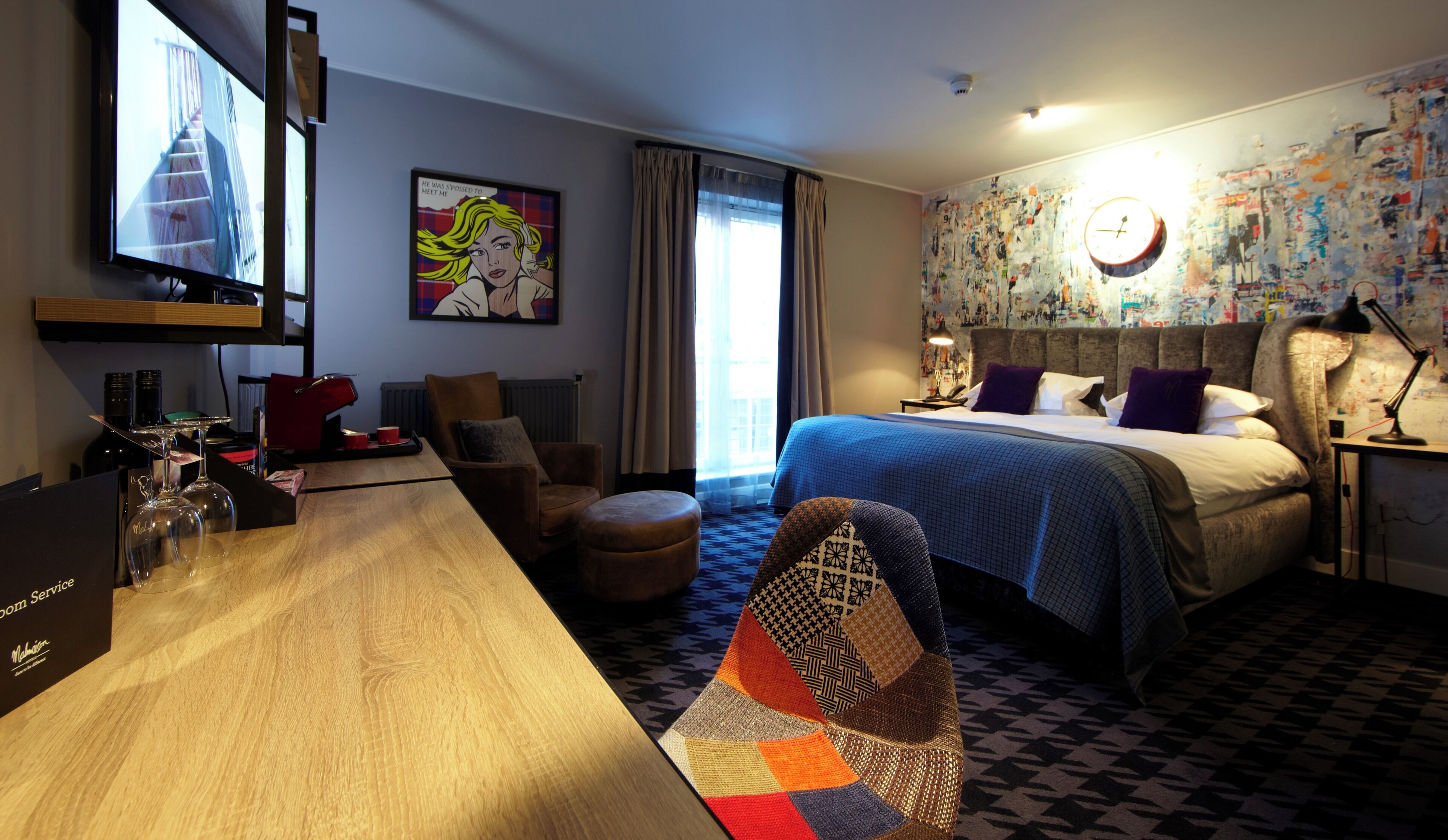 Camera d'albergo con un letto, una scrivania con televisore, una sedia, un tavolino con bicchieri da vino e un tappeto colorato.
