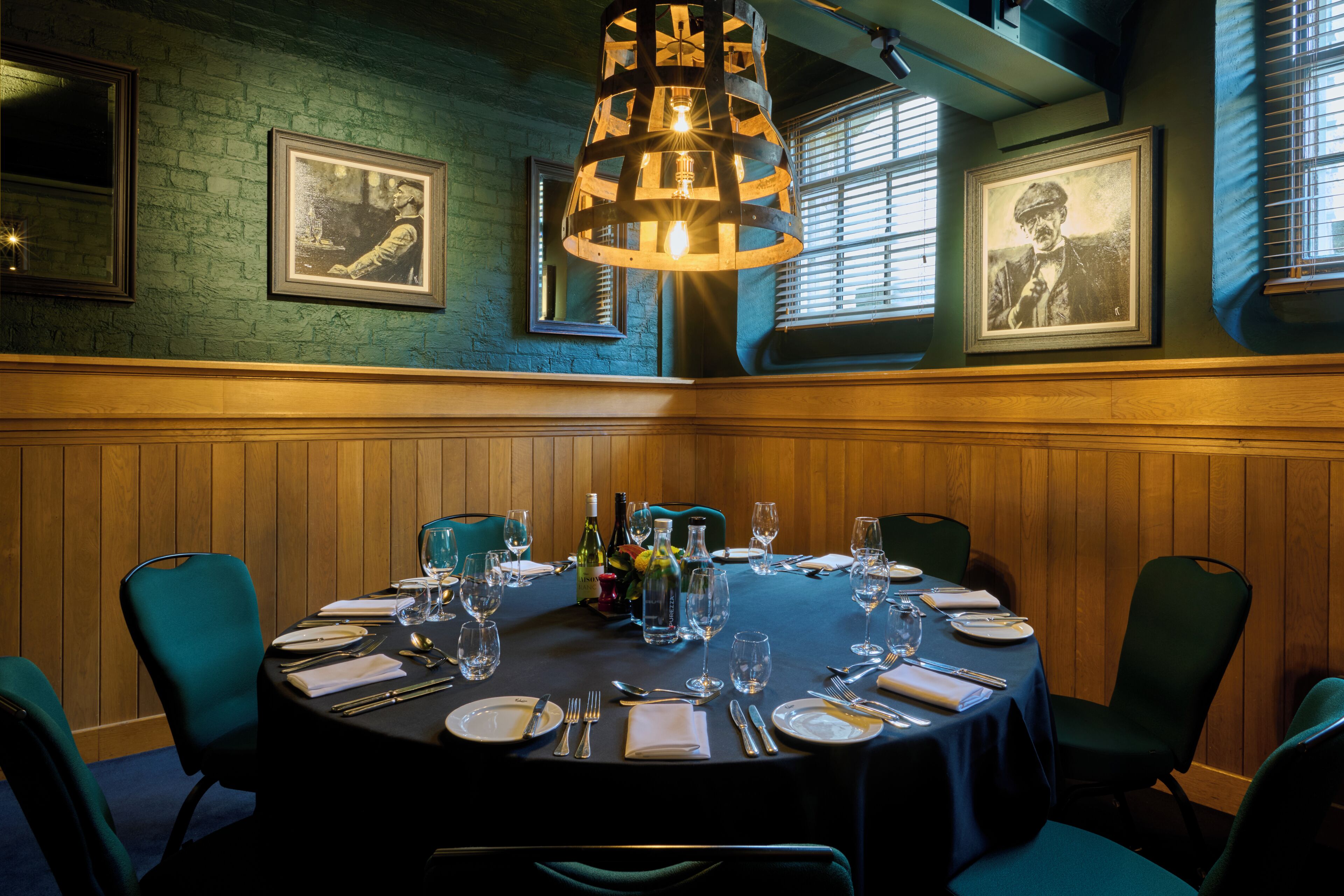 A dining room with a round table set for six, wooden paneled walls, framed pictures, and a hanging light fixture.