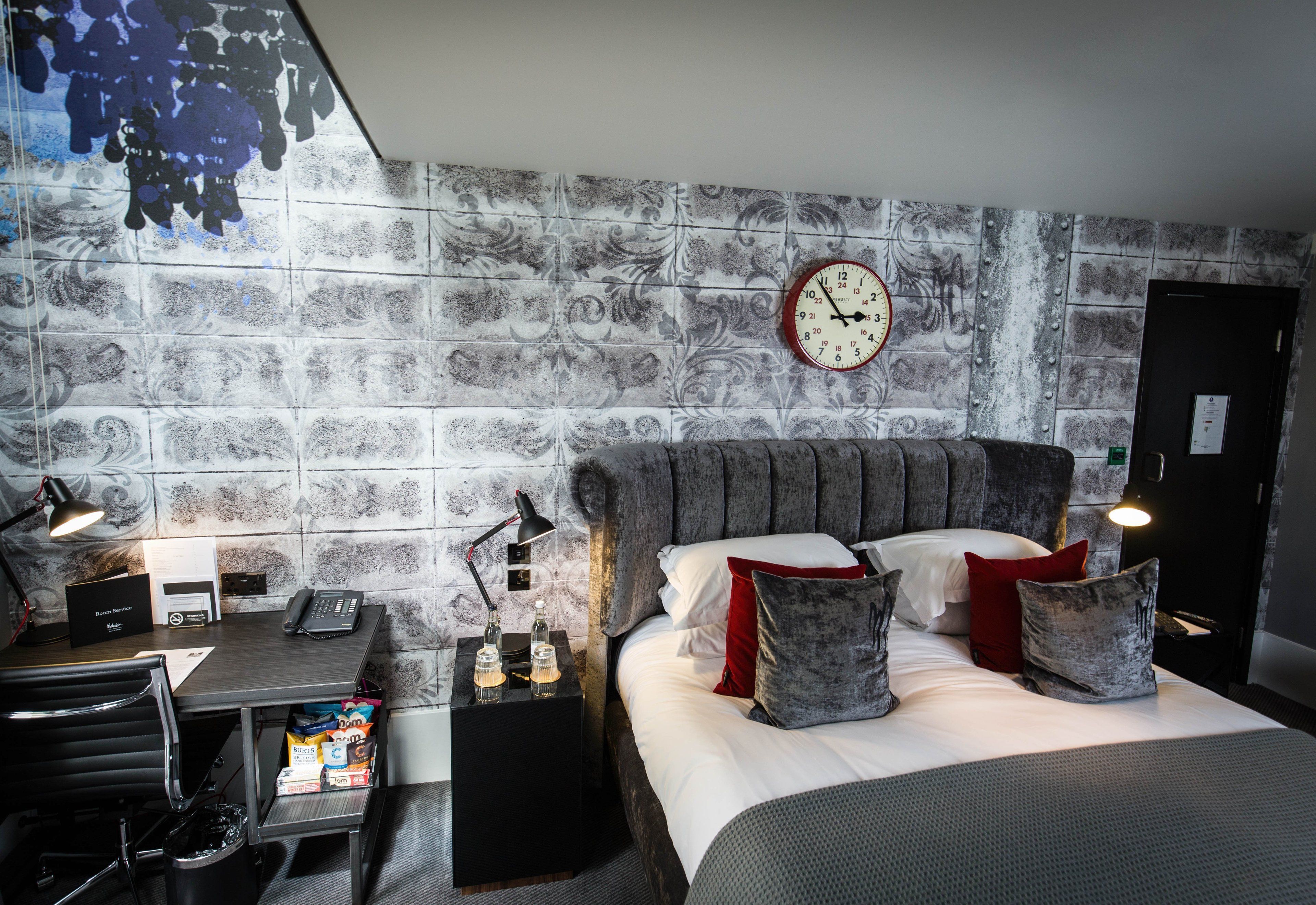 A hotel room with a bed, a desk with a telephone, a chair, a trash bin, and a wall clock.