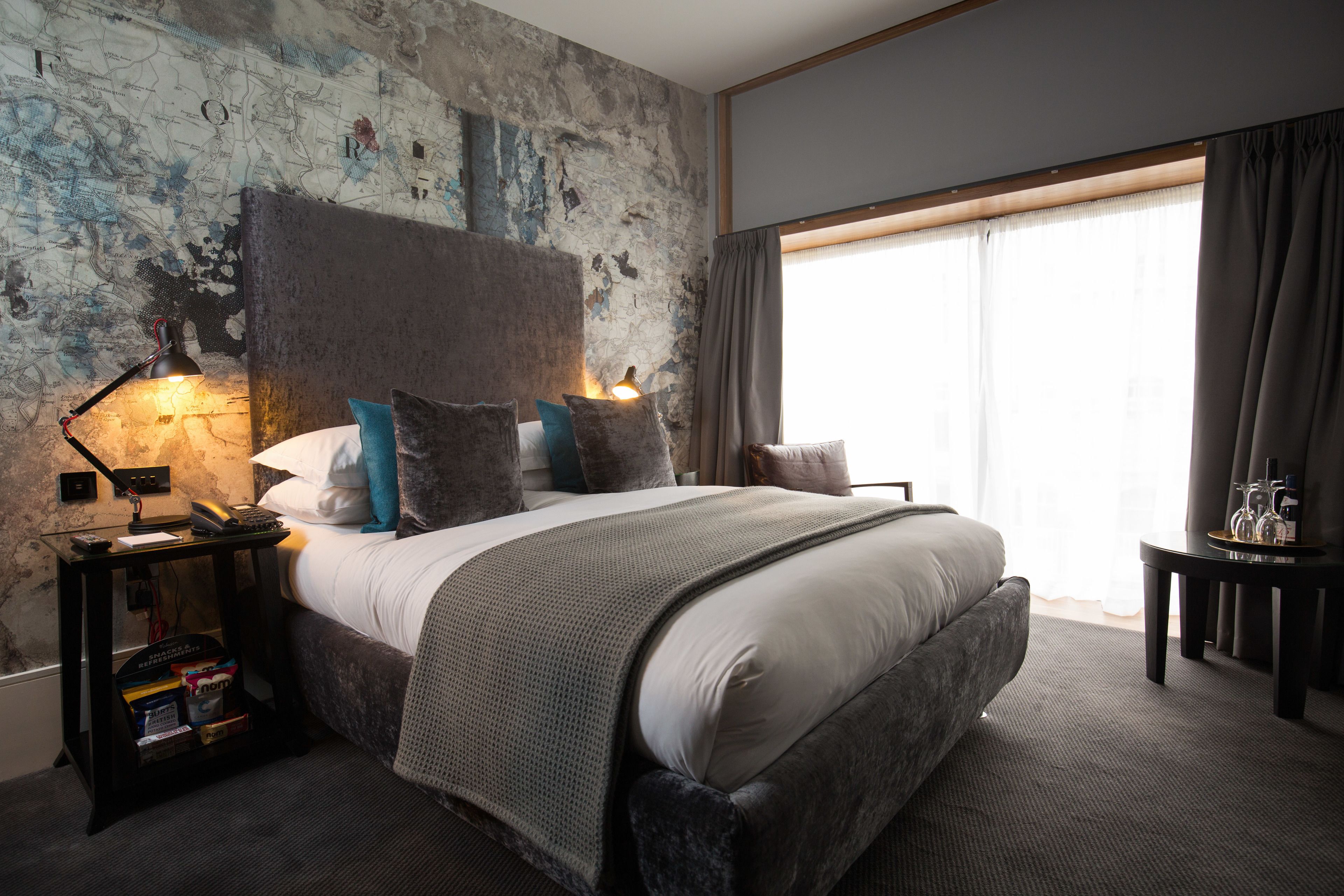 A modern hotel room with a large bed, a nightstand with a lamp, a small table with a tray, and a view of the outdoors through a large window.