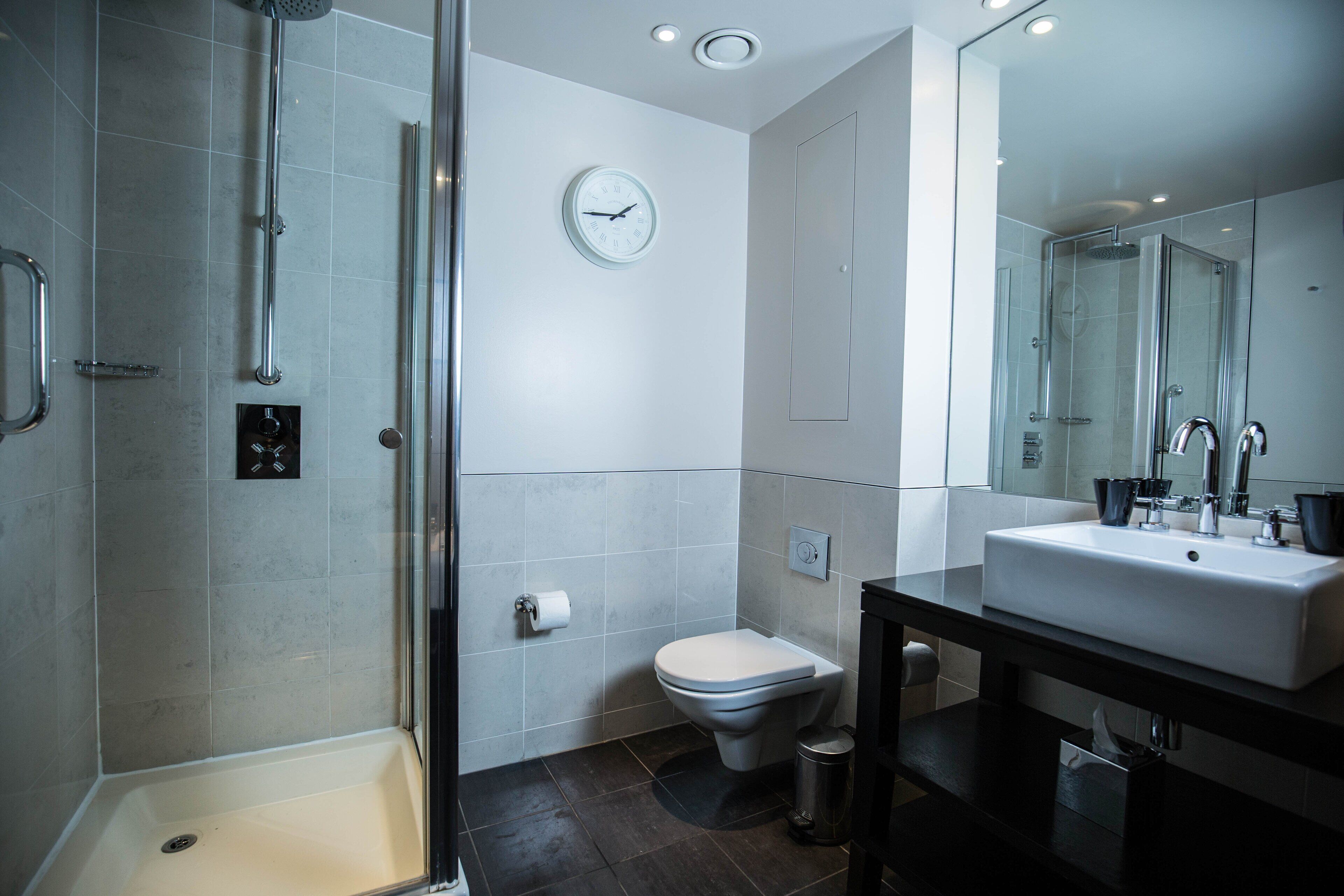 A modern bathroom with a glass shower enclosure, a toilet, a sink with a mirror, and a wall clock.