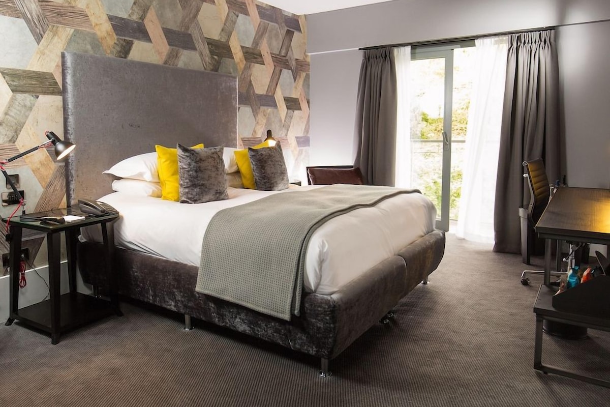 A modern hotel room with a large bed, a desk, and a view of greenery through the window.