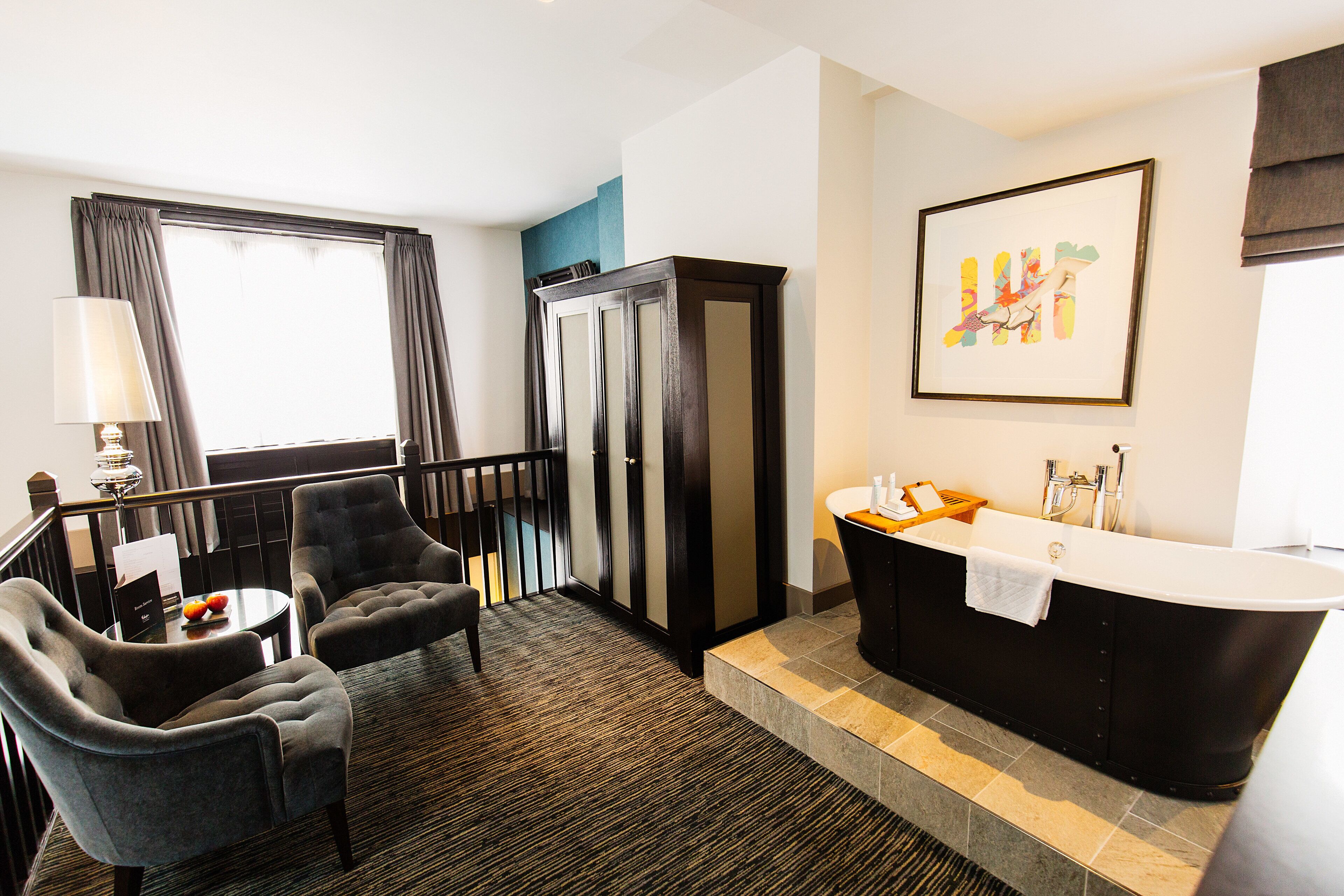 A hotel room with a black bathtub, two armchairs, a small table with fruit, a lamp, and a window with curtains.