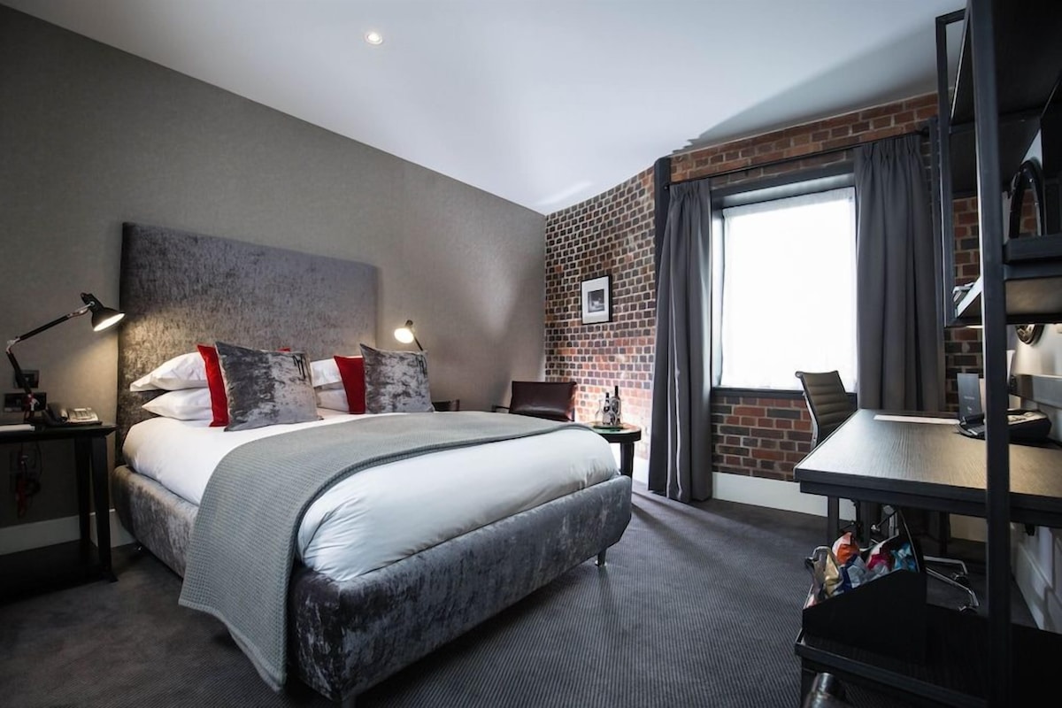 A hotel room with a bed, a desk, a chair, a lamp, a window with curtains, and a brick wall.